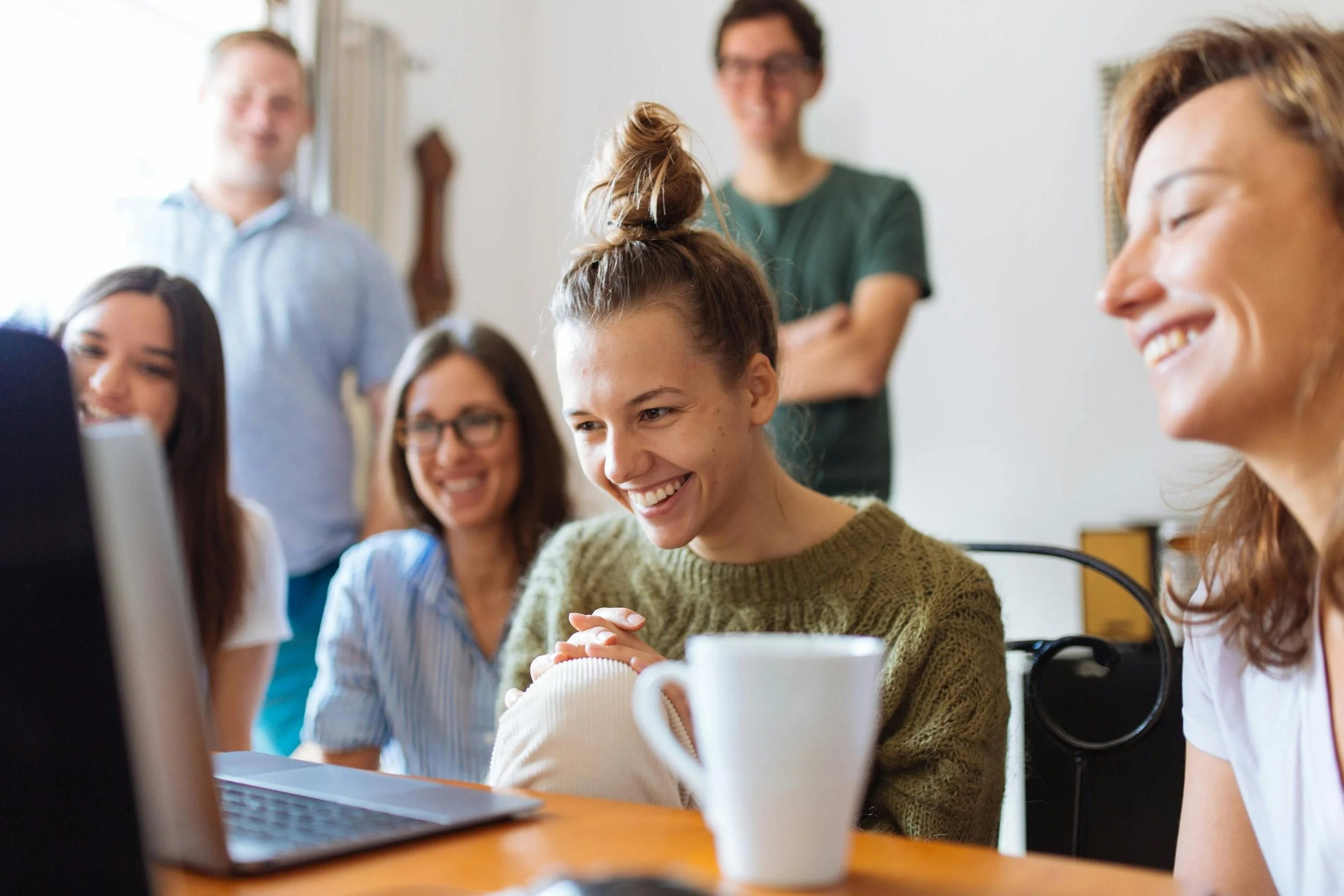 Six people smiling and looking at a laptop in a casual office or home setting.