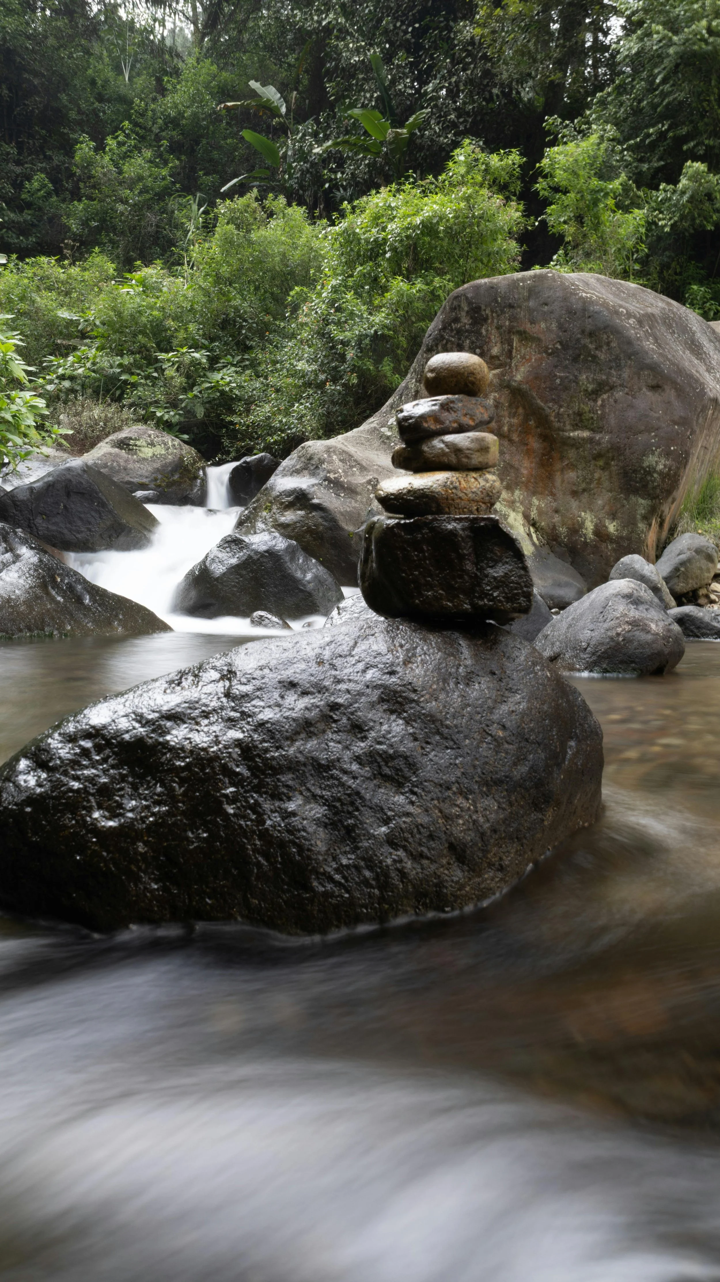 A natural stream flowing over rocks, with a stack of small stones balanced on a large rock in the foreground, surrounded by lush green foliage.