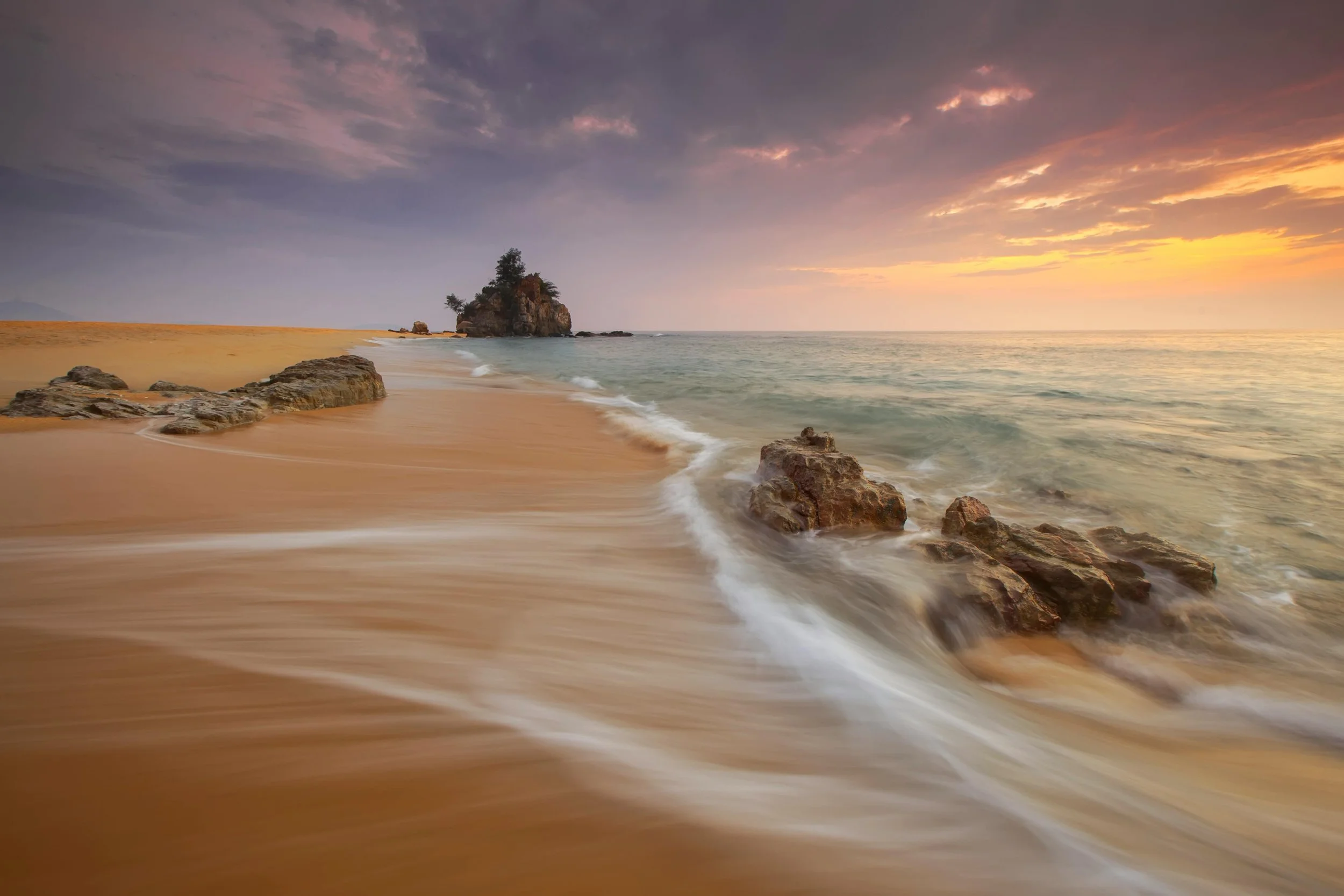 Sunset over a beach with rocks and an island with trees in the distance, gentle waves washing onto the sandy shore.