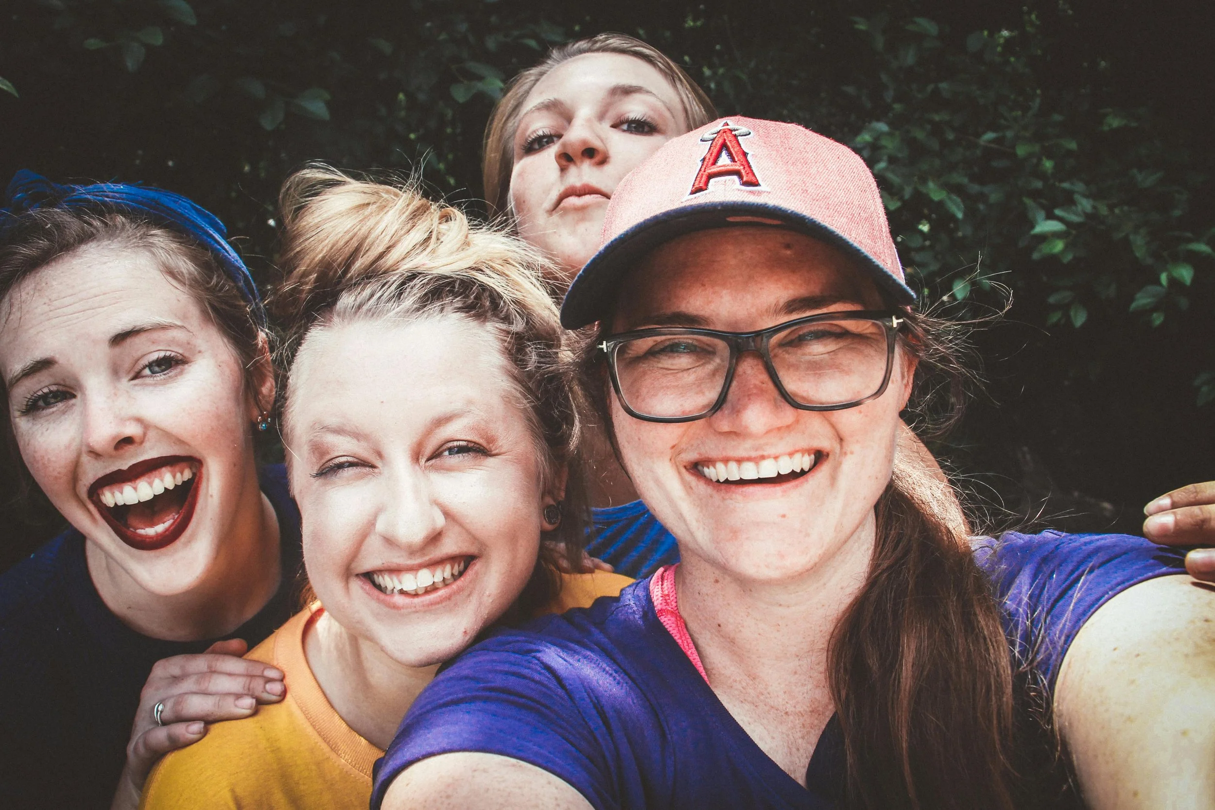 Five women smiling and taking a group selfie outdoors with greenery in the background.