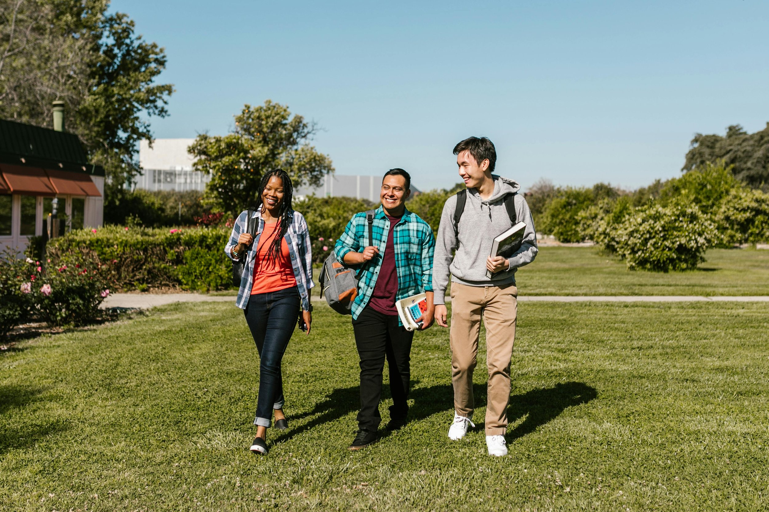 Three young adults walking outdoors on a sunny day, carrying books and backpacks, smiling and talking.