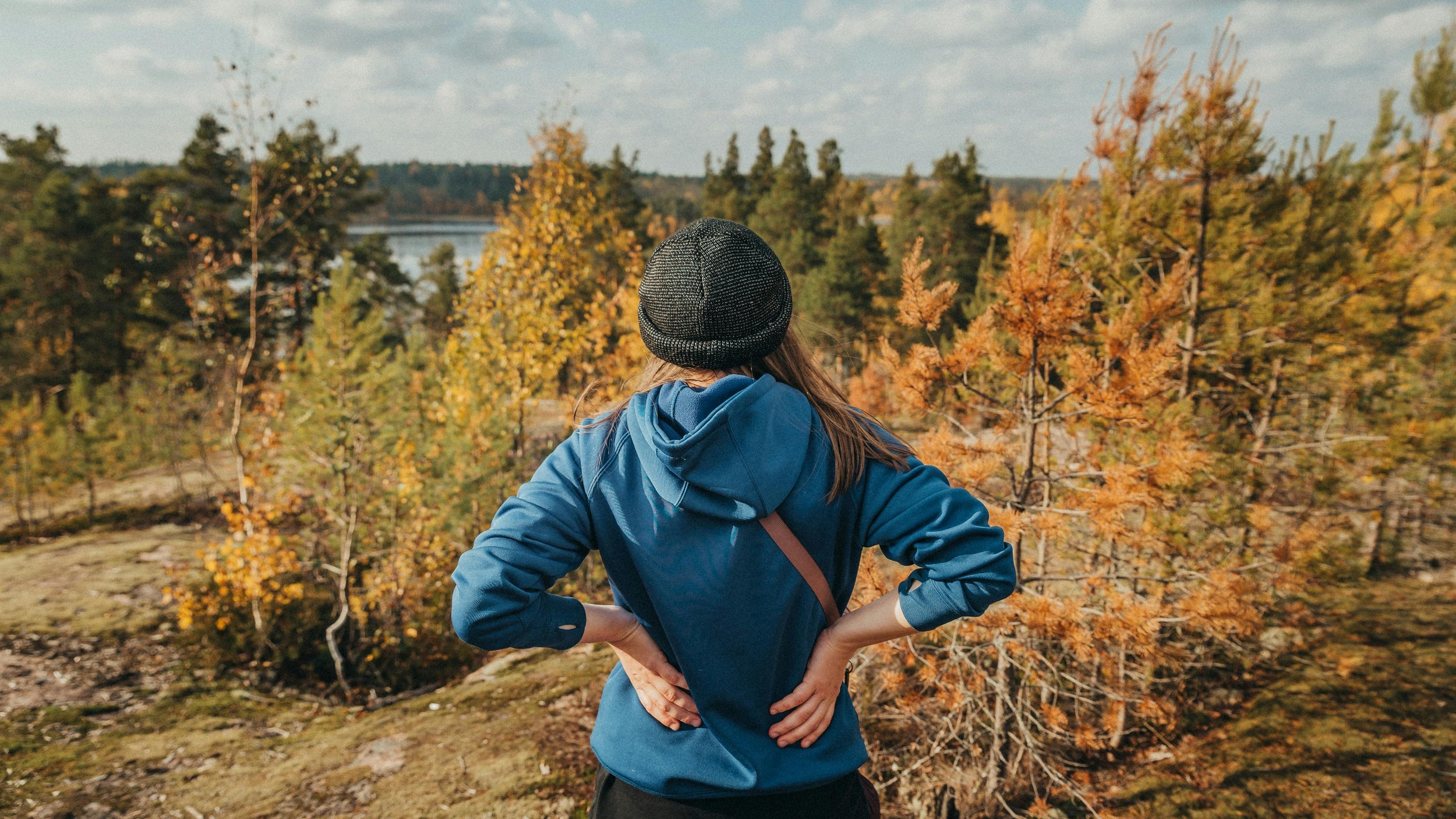 Person in a blue jacket and black beanie looking at fall foliage near a body of water.