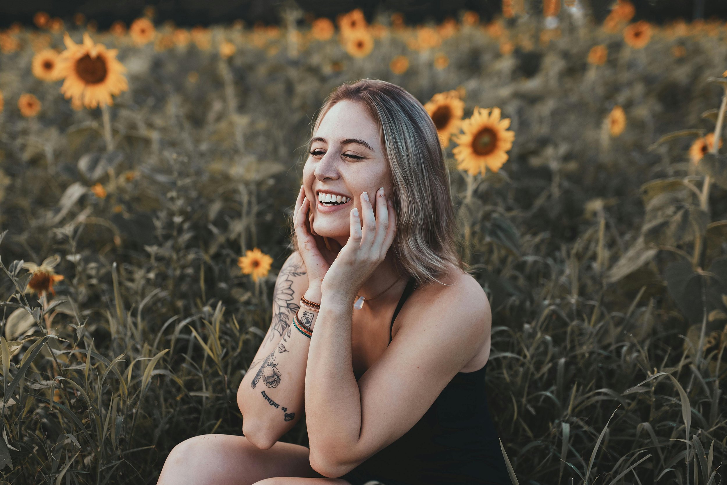A woman sitting in a sunflower field, smiling with her hands on her face.