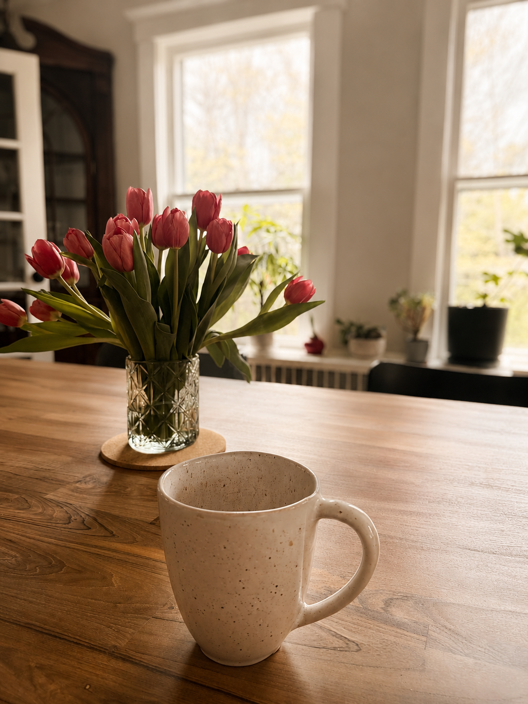 A beige ceramic mug on a wooden table, with a vase of pink tulips in the background and sunlight coming through large windows.