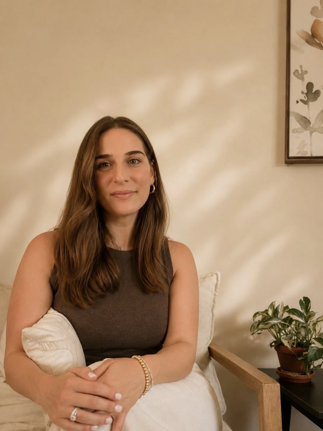 A woman with long brown hair sitting on a beige couch, holding a white pillow, wearing a dark sleeveless top and jewelry, in a cozy room with cream-colored walls, a framed botanical print, and a potted plant on a side table.
