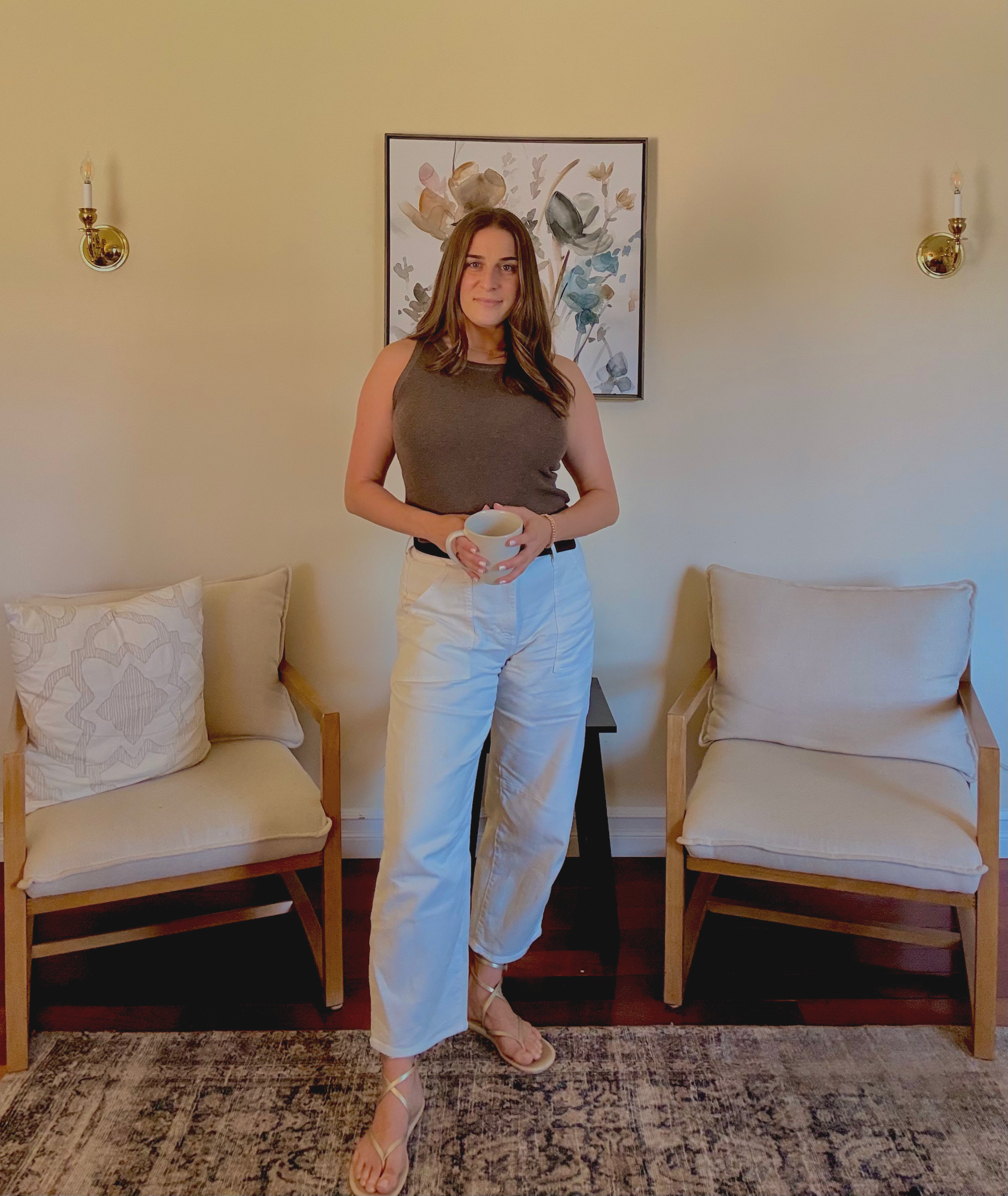 A woman with medium-length brown hair standing in a living room, holding a white mug, wearing a sleeveless brown top, white pants, and strappy sandals, with two beige chairs and a floral painting on the wall behind her.