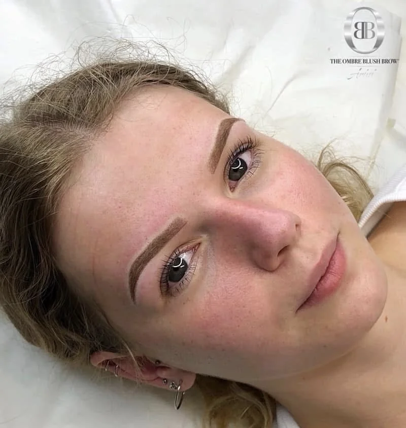 Close-up of a young woman lying down with freshly done eyebrow makeup or microblading, showcasing well-defined eyebrows, with a slight smile, curly blonde hair, and multiple earrings in her ear.
