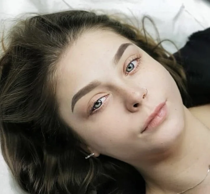Close-up of a young woman with fair skin, dark wavy hair, light-colored eyes, and well-defined eyebrows lying down, looking at the camera.