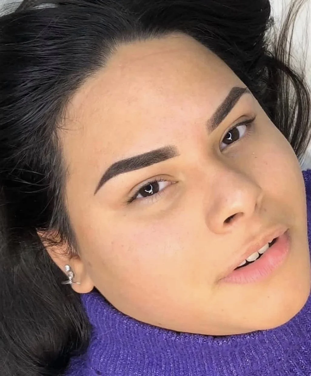 A close-up of a woman's face featuring well-groomed eyebrows, dark hair, and a purple top.