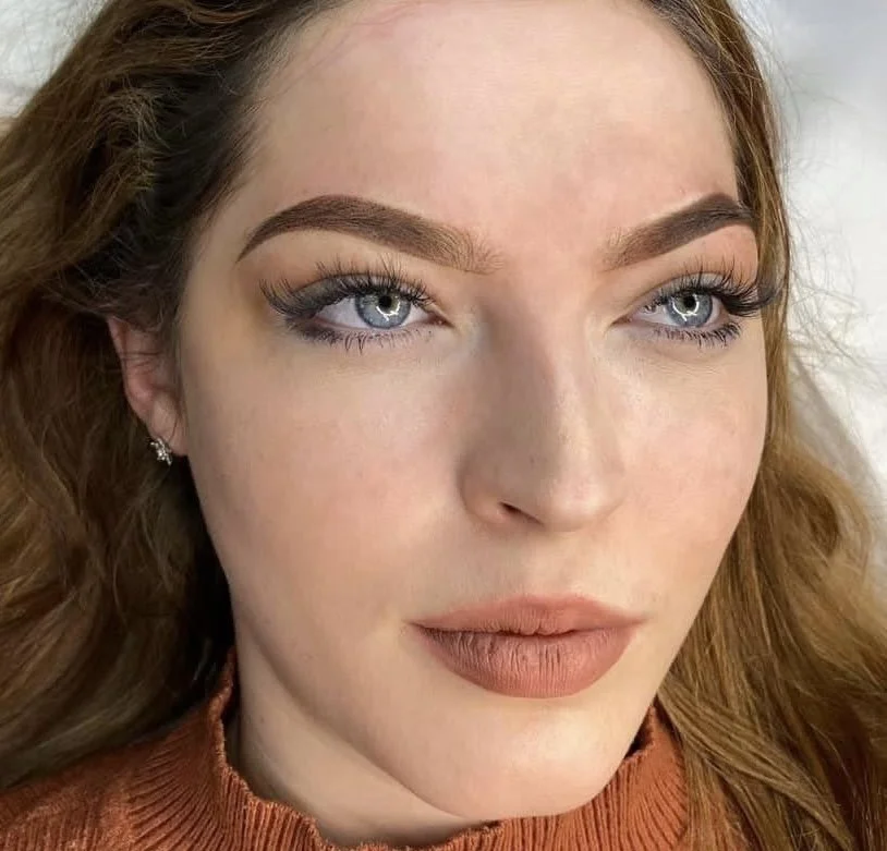 Close-up of a woman with blue eyes, brown eyebrows, and long eyelashes, wearing makeup with nude lipstick, and a rust-colored top.