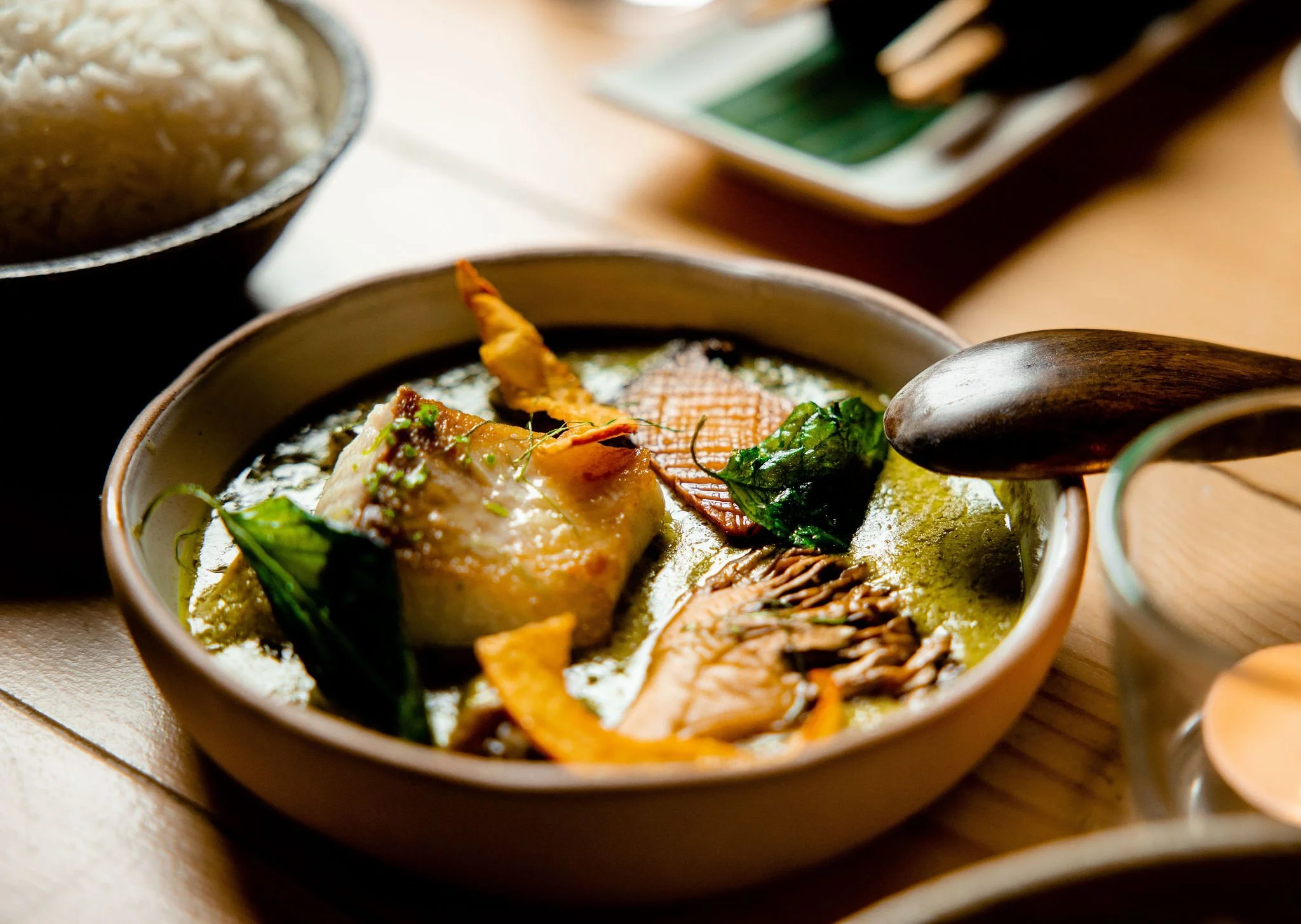 Bowl of green curry with chicken, vegetables, basil, and crispy toppings, served with a wooden spoon and a side of white rice.