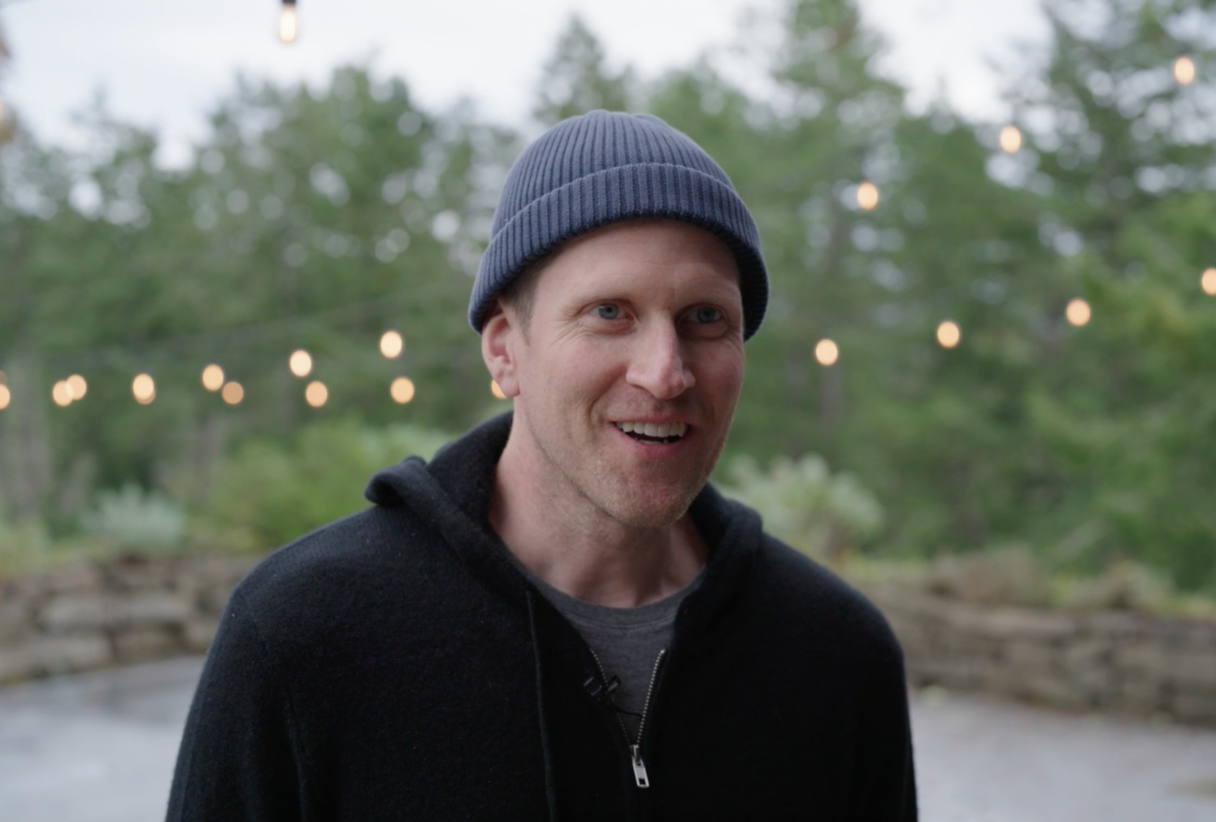 A smiling man with light skin, short hair, wearing a navy blue beanie and black zip-up hoodie outdoors with string lights and green trees in the background.