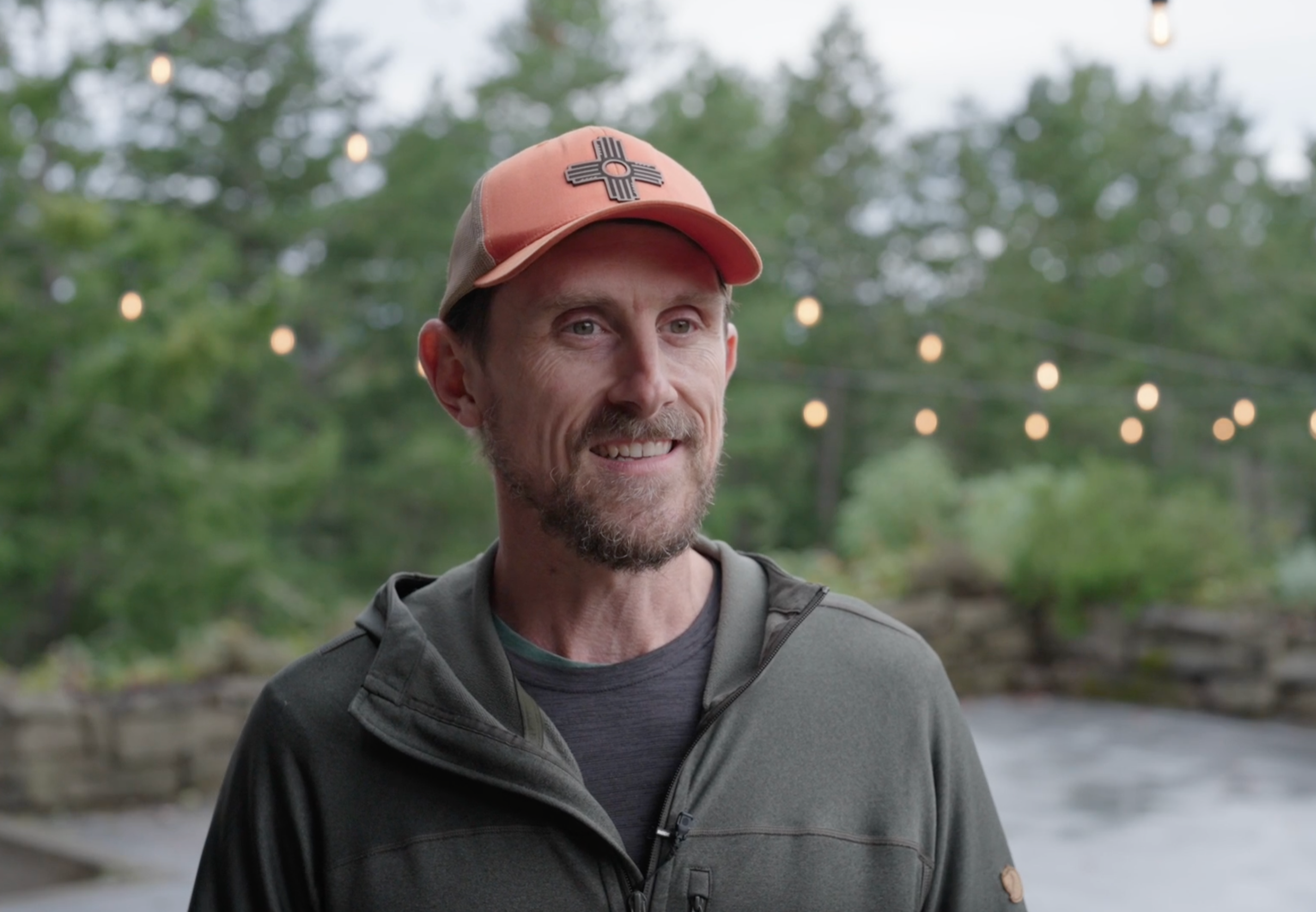 A smiling man wearing a cap and outdoor jacket, standing outdoors with a blurred background of trees and string lights.