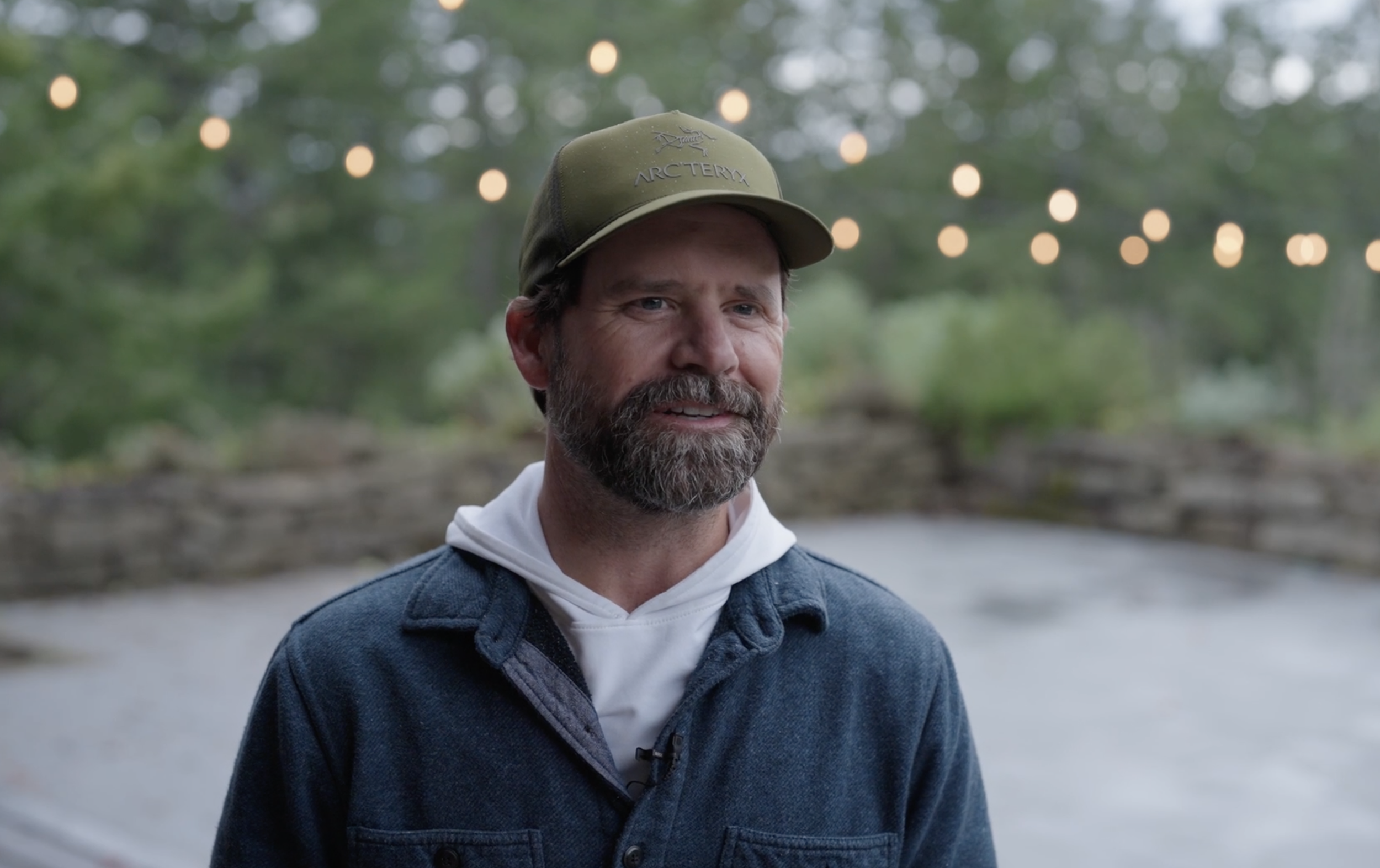 A man with a beard wearing a cap and jacket, standing outdoors with string lights and trees in the background.