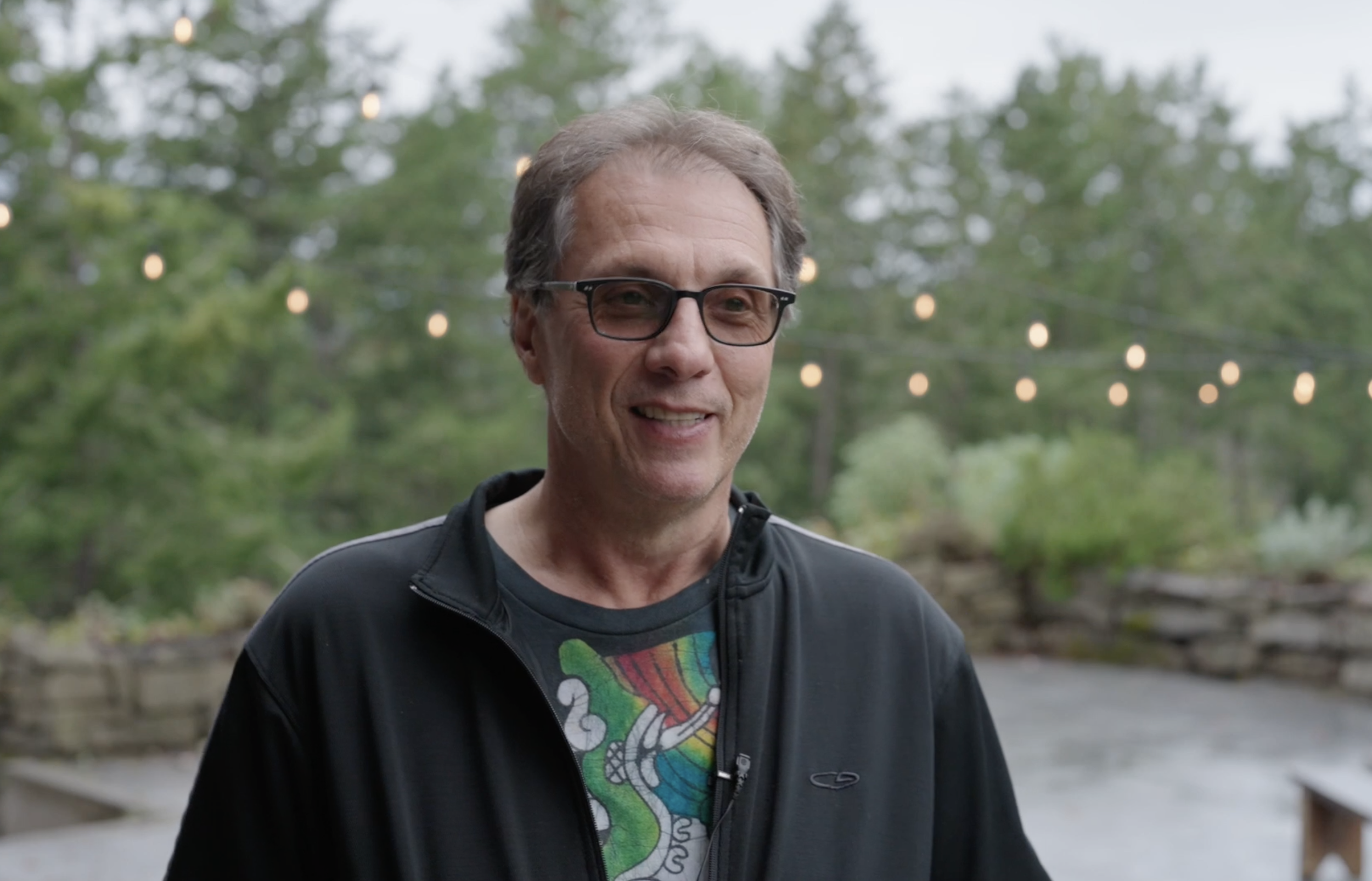 A middle-aged man with glasses smiling outdoors with greenery in the background and string lights overhead.
