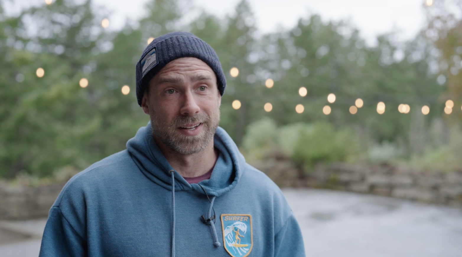 Man with a gray beard wearing a blue hoodie with a surfing patch, a navy knit beanie, and a lavalier microphone, outdoors with string lights and greenery in the background.
