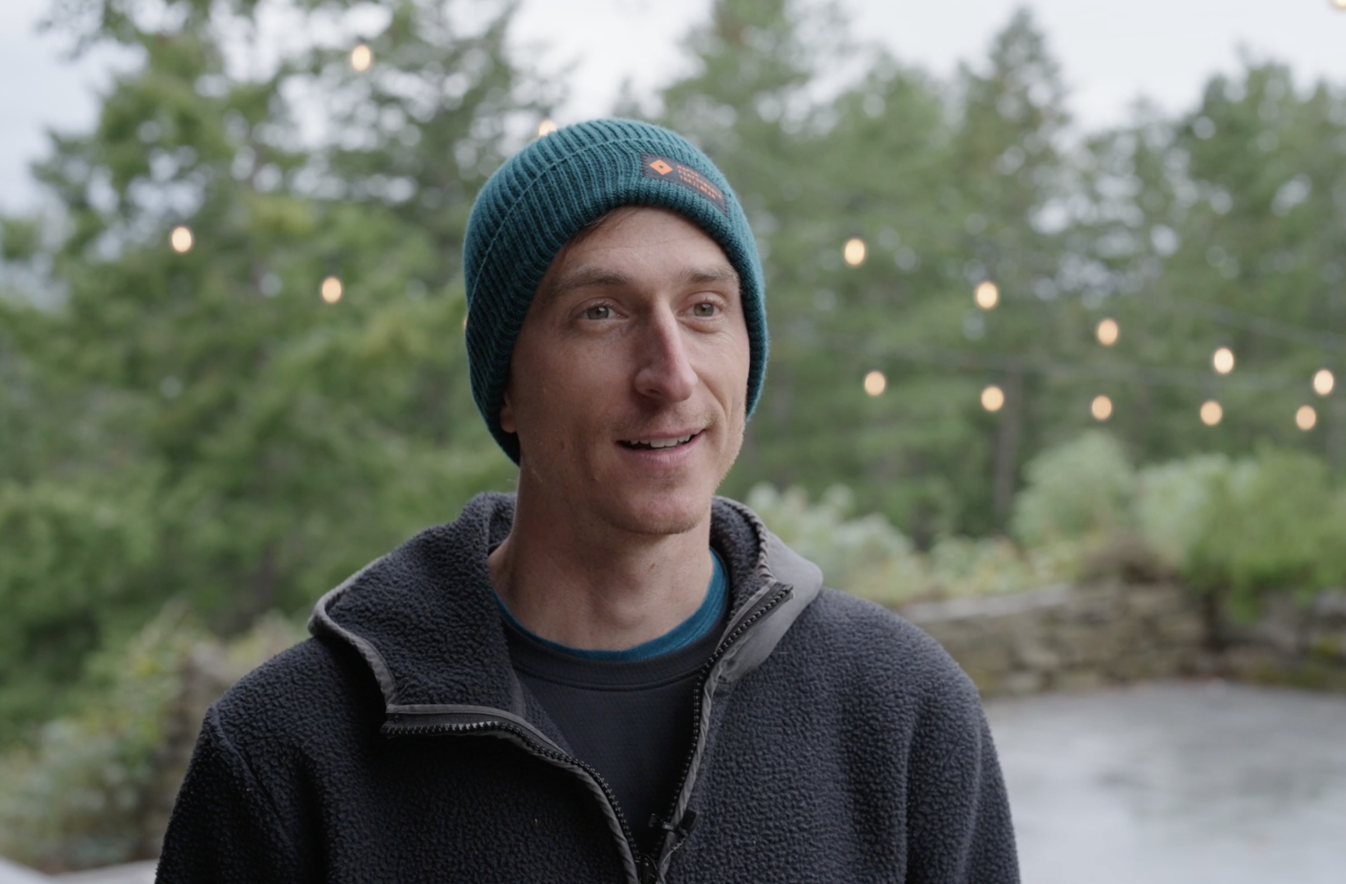 A man wearing a teal beanie and black fleece jacket, smiling outdoors with blurred trees and string lights in the background.