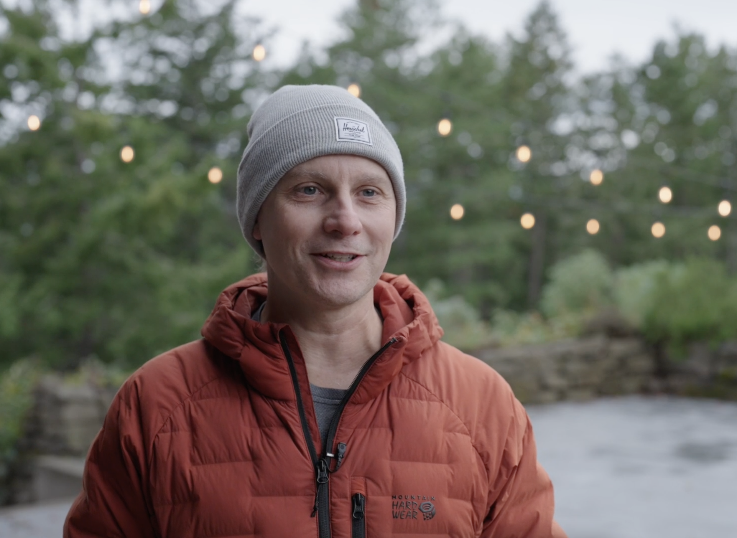 A man wearing a gray beanie and an orange jacket outdoors, with string lights and green trees in the background.