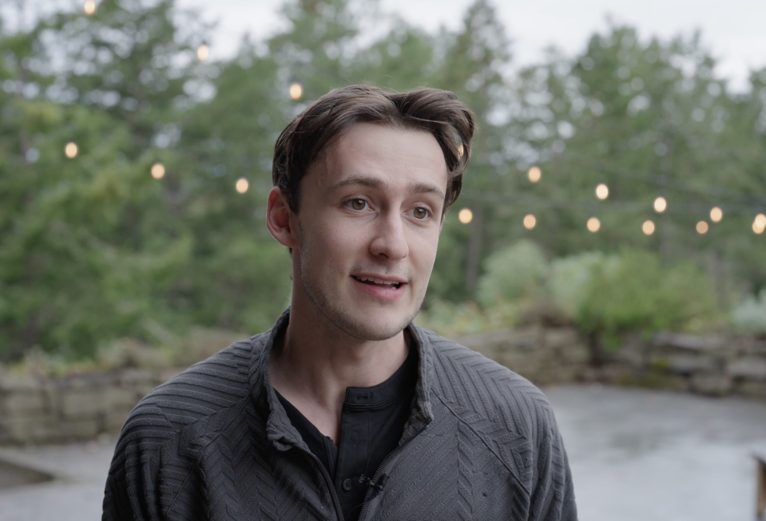A young man with brown hair talking outdoors with string lights and green trees in the background.