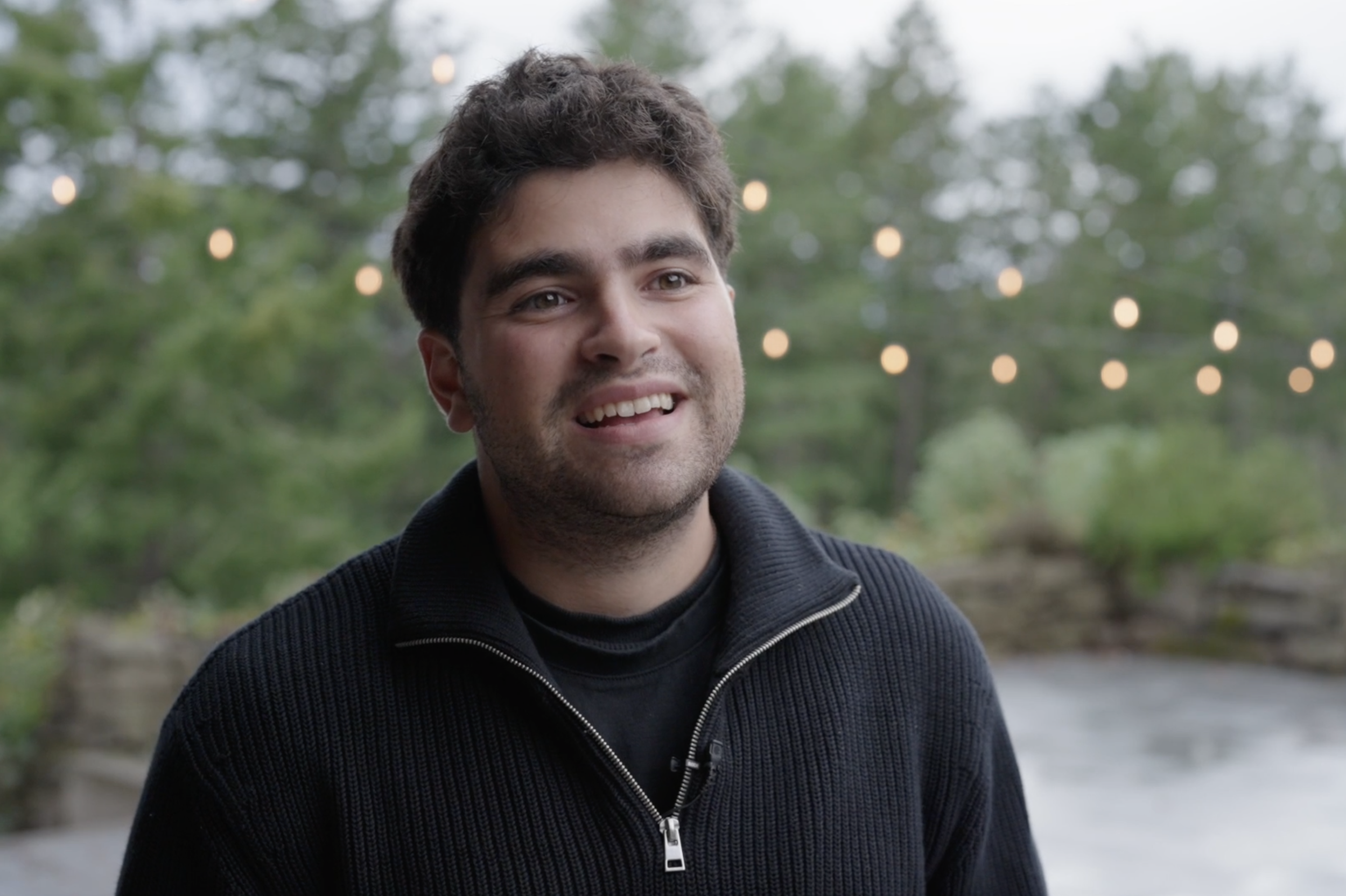 A smiling man with dark hair and beard, wearing a black zip-up sweater, standing outdoors with blurry trees and string lights in the background.