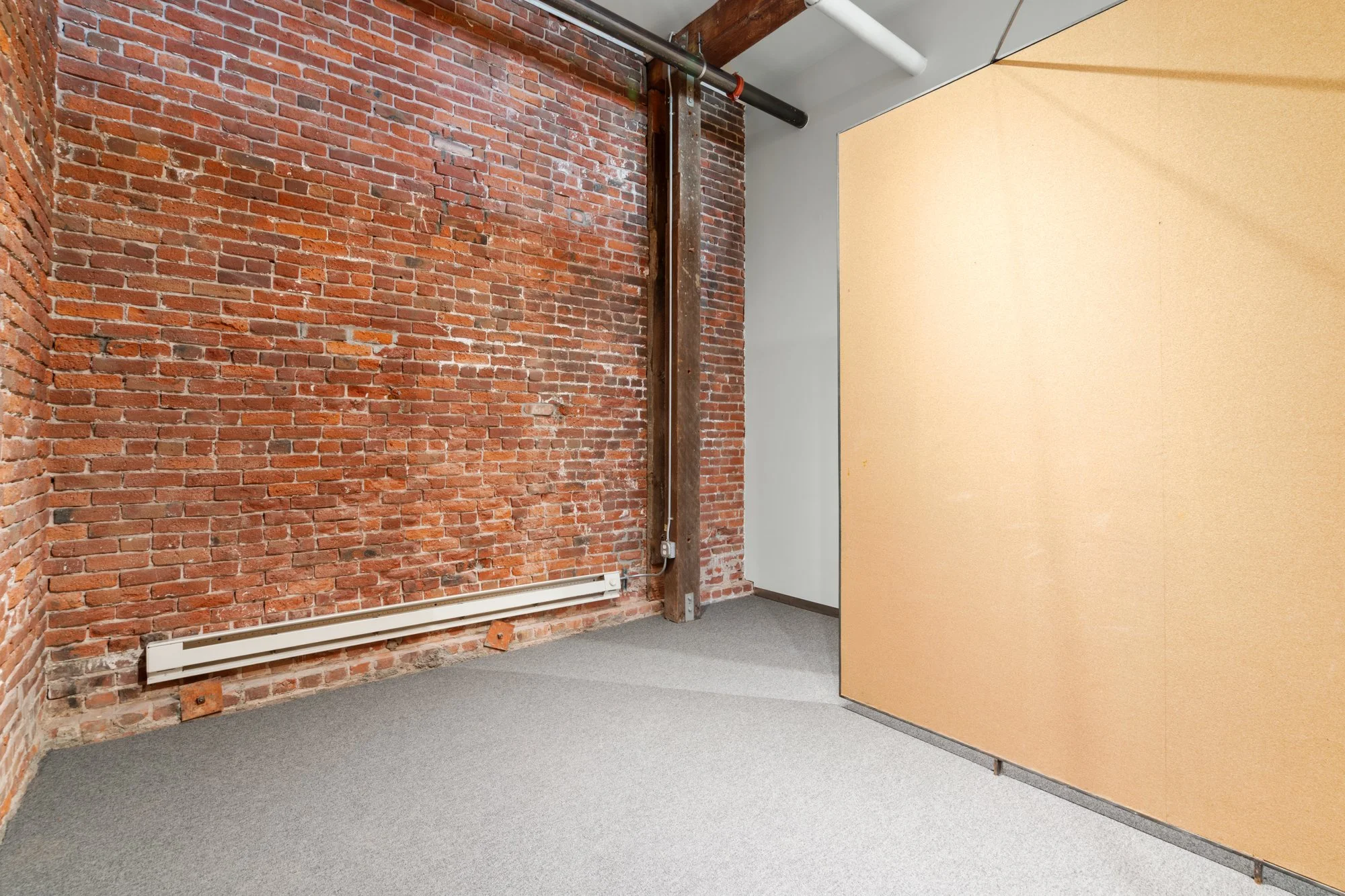 Empty room with red brick wall, white baseboard heater, exposed metal pipe, and a partition wall on the right.