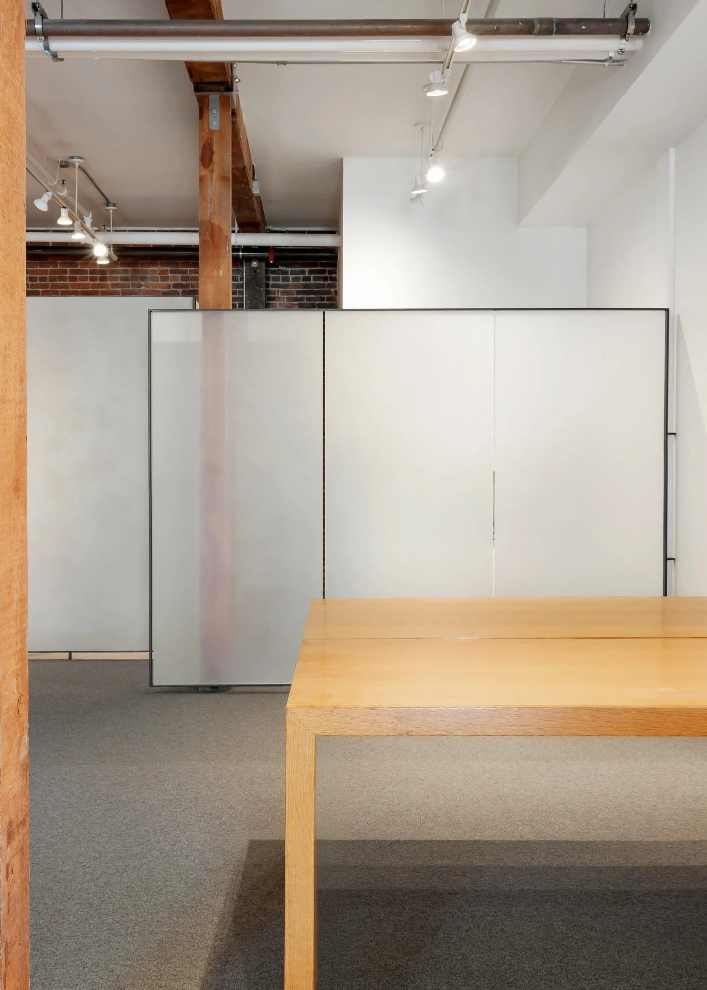 Empty room with white partitions, a wooden table in the foreground, and exposed brick and wood beams on the ceiling.