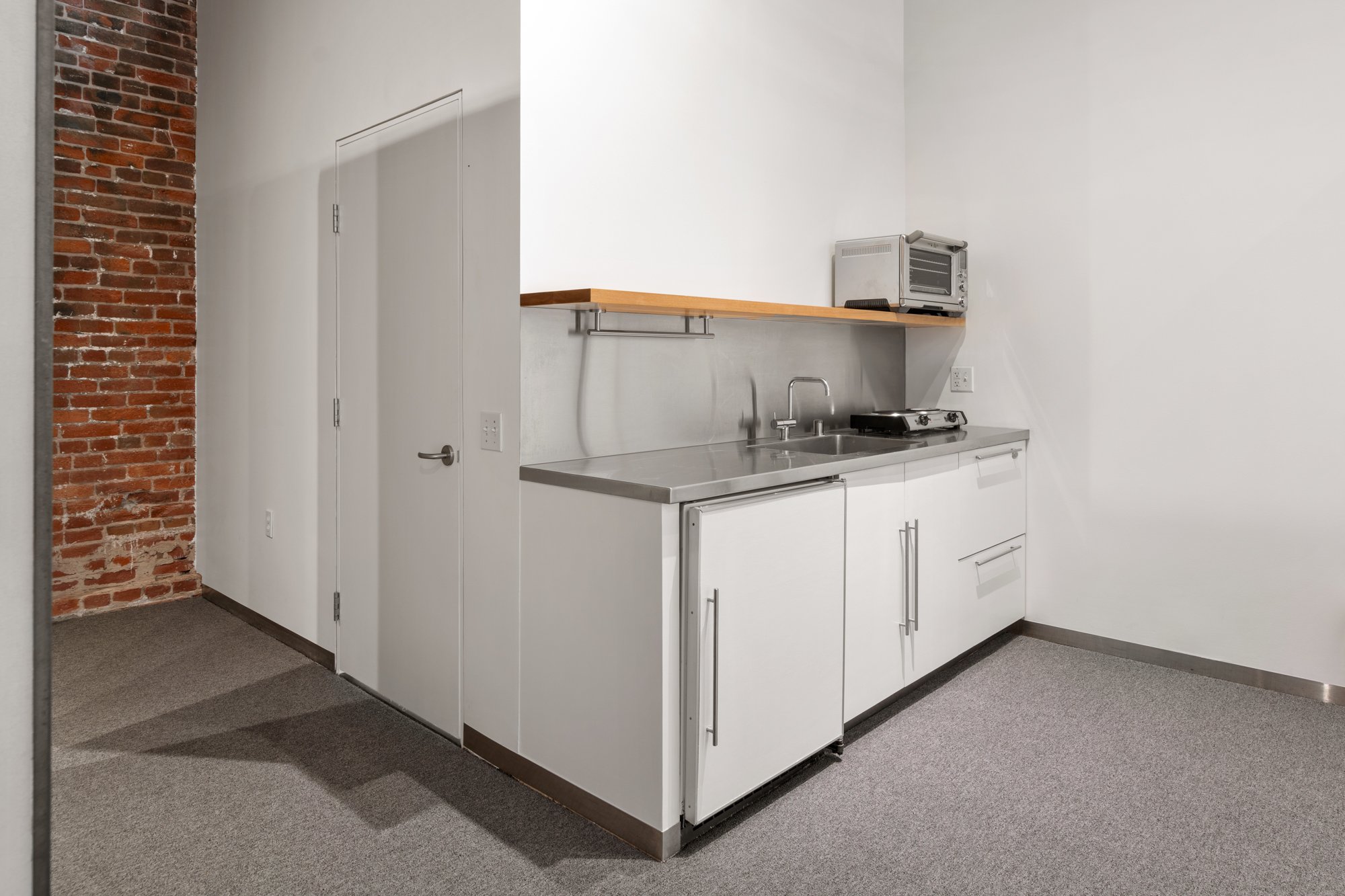 A small kitchenette with white cabinets, a stainless steel sink, and a two-burner stove. There is a wooden shelf above the counter with a toaster oven on it. The wall behind the sink and stove is white, and there is an exposed brick wall on the left 
