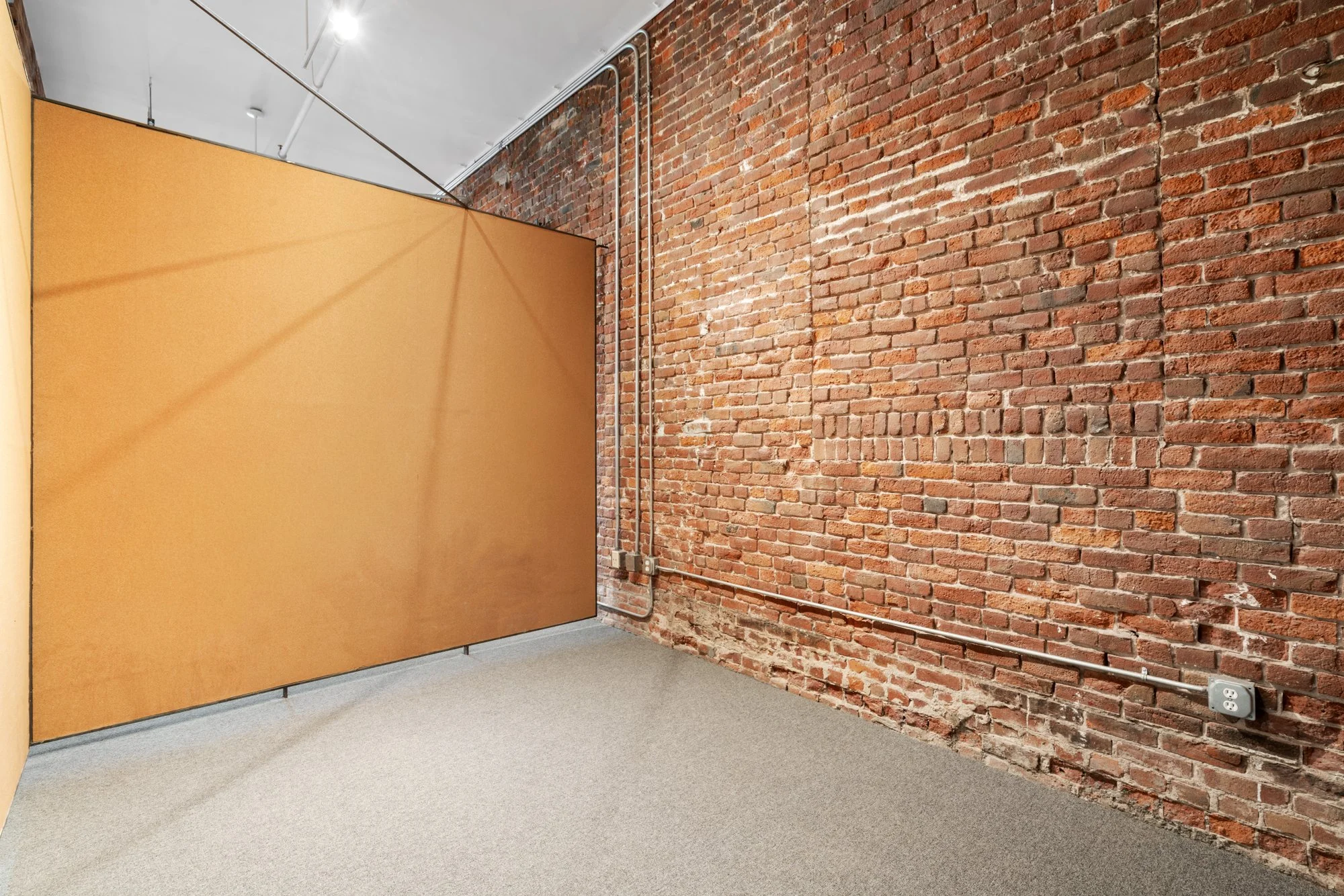 Empty office corner with exposed brick wall, gray carpet, and a partial orange partition.