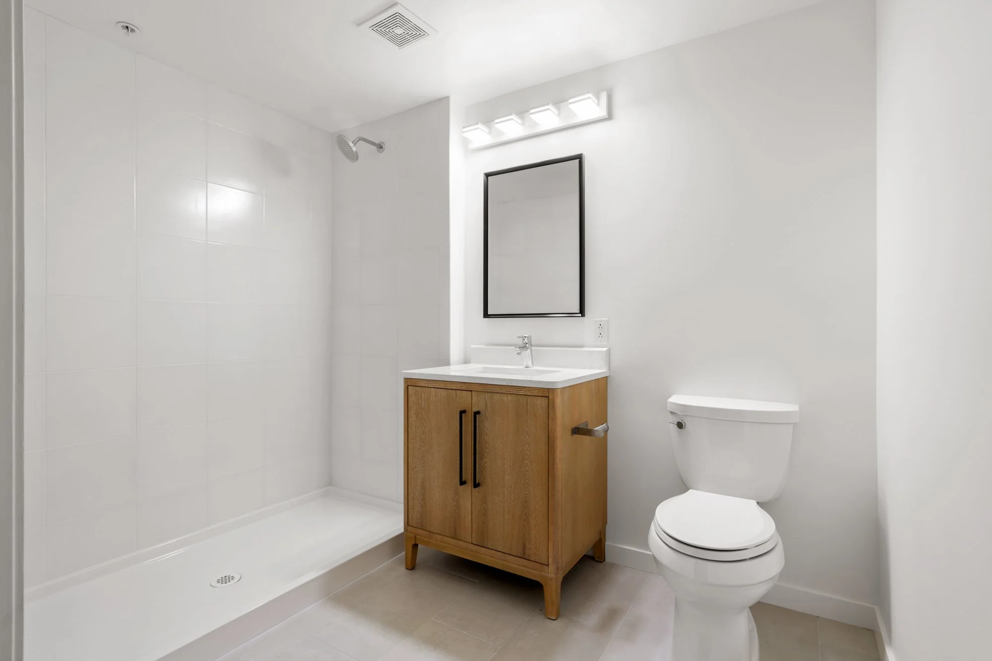 Modern bathroom with a walk-in shower, wooden vanity with a white sink, black-framed mirror, wall-mounted light fixture, and a white toilet.