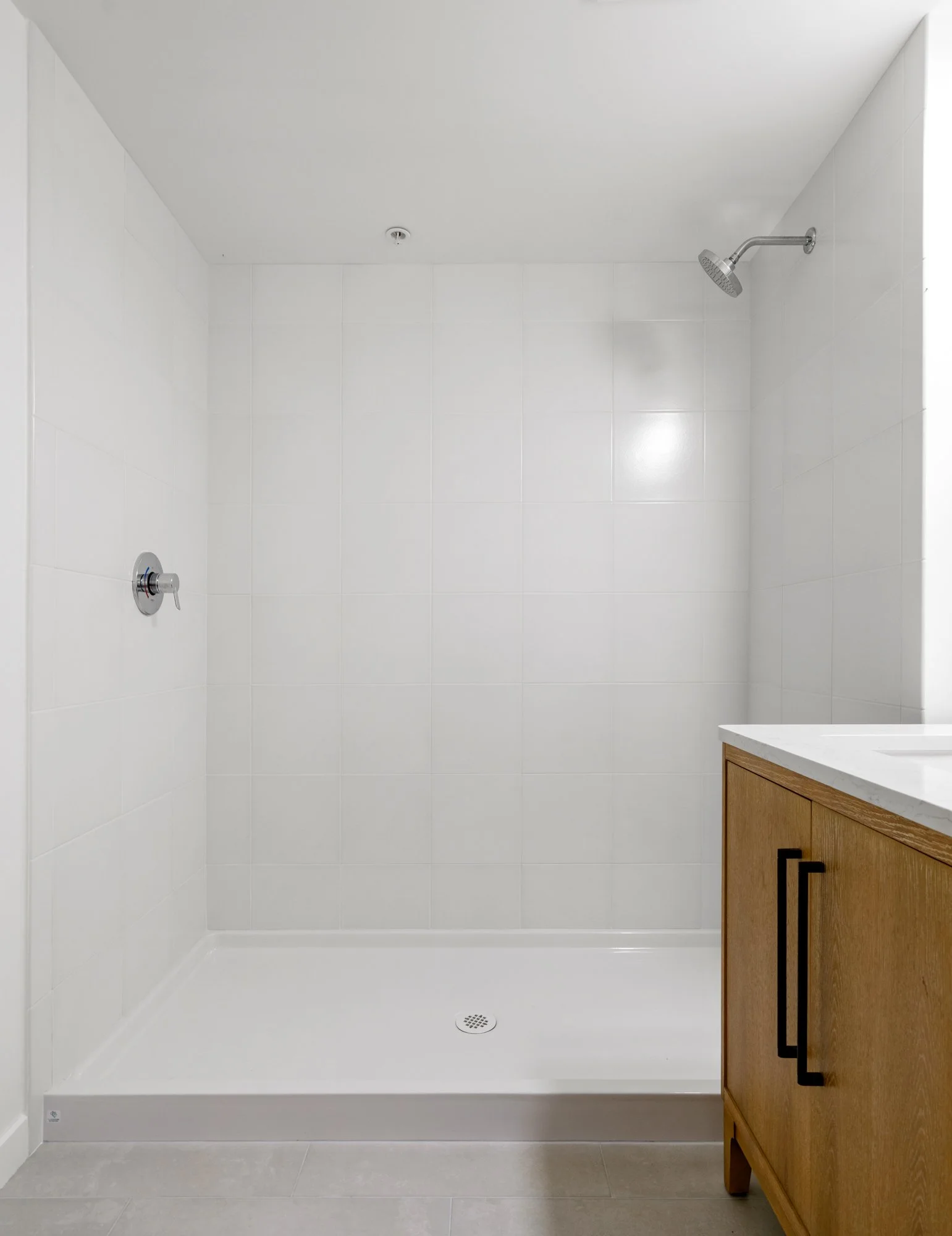 Empty white shower with a drain on the floor and a wooden bathroom cabinet with black handles on the right.