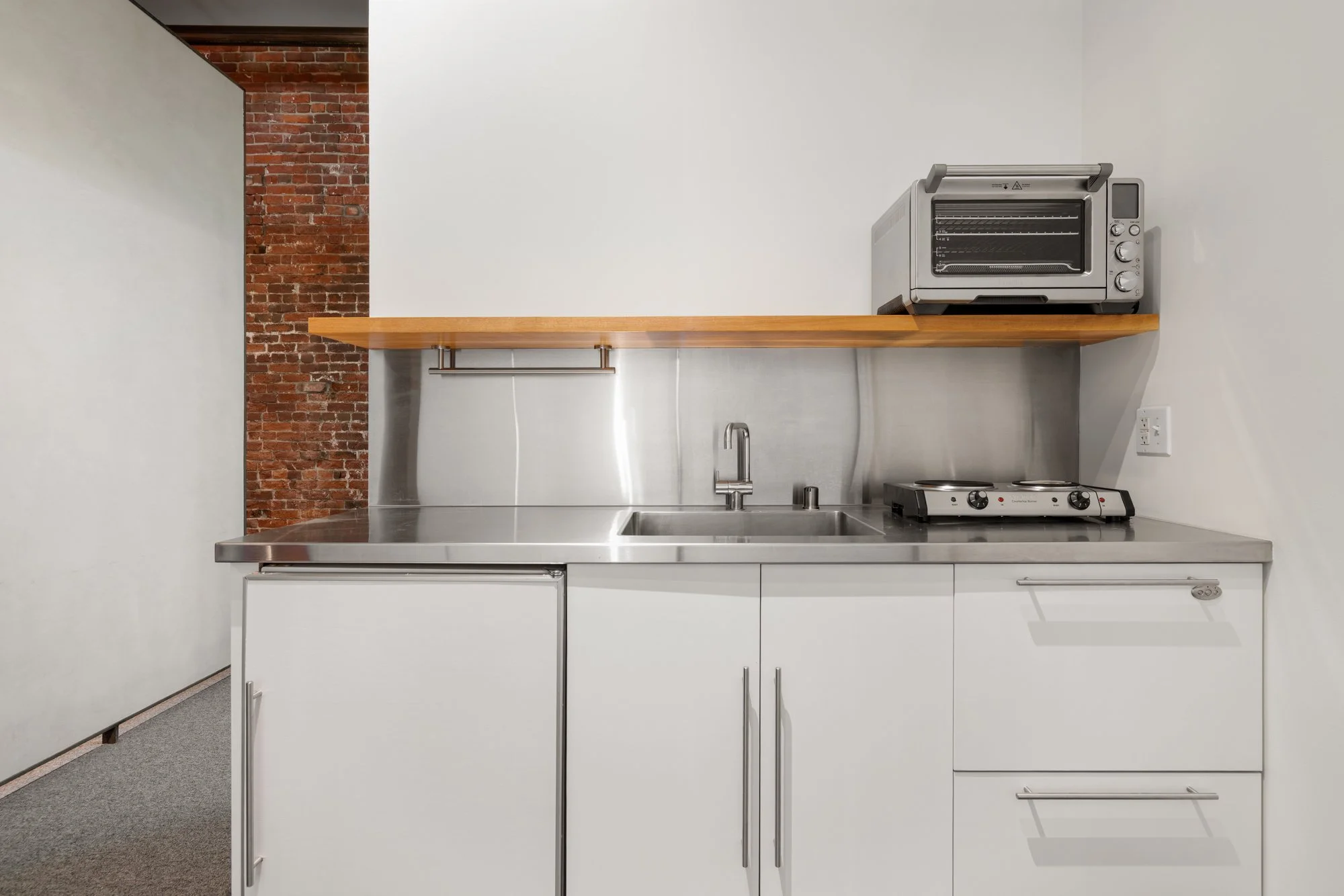 Small kitchen with white cabinets, a stainless steel countertop, a sink, a two-burner electric stove, a microwave oven on a wooden shelf, and a partial view of a brick wall.