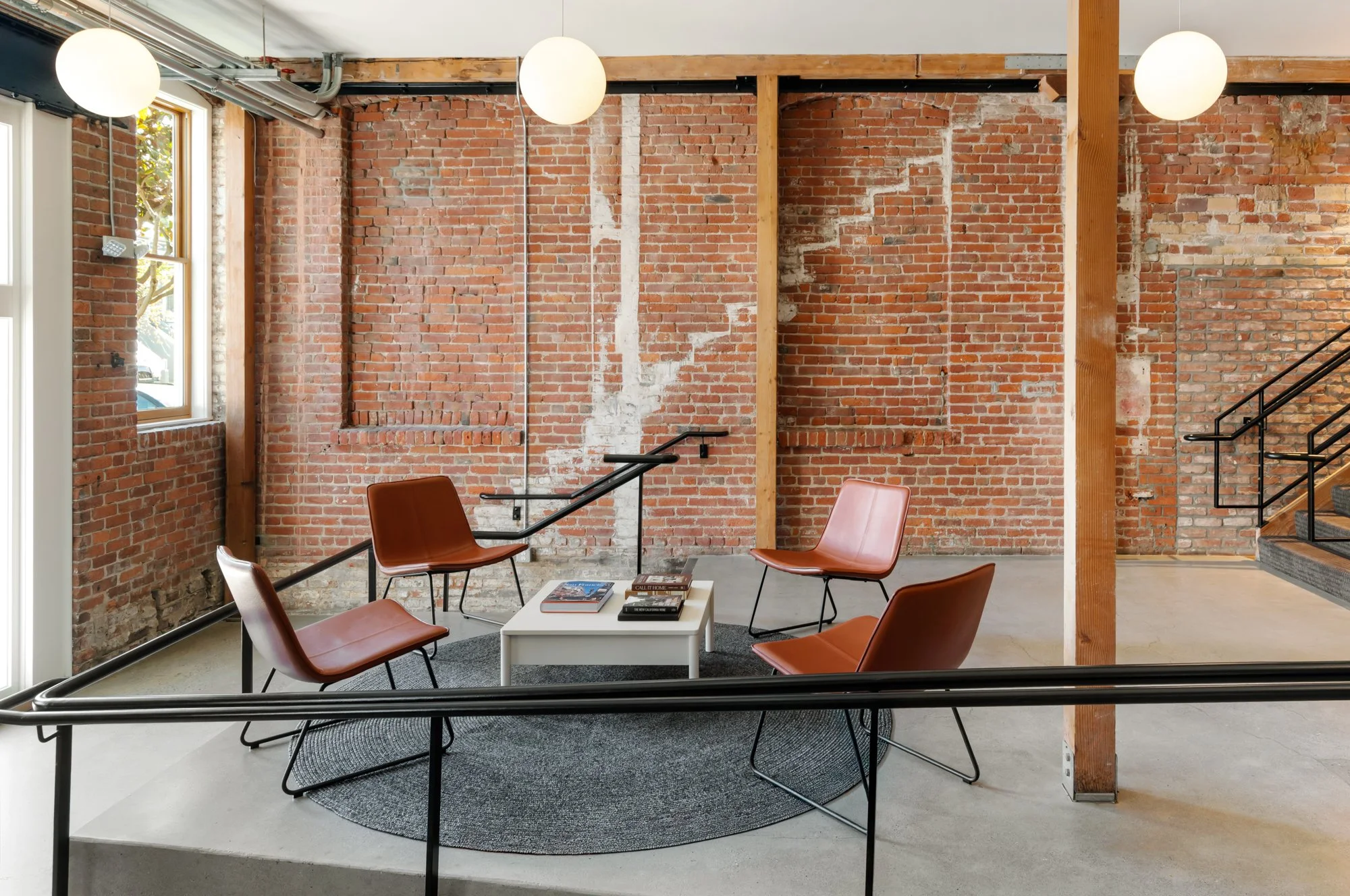 Modern interior with exposed brick walls, four brown chairs around a white coffee table on a round black rug, and large windows letting in natural light.