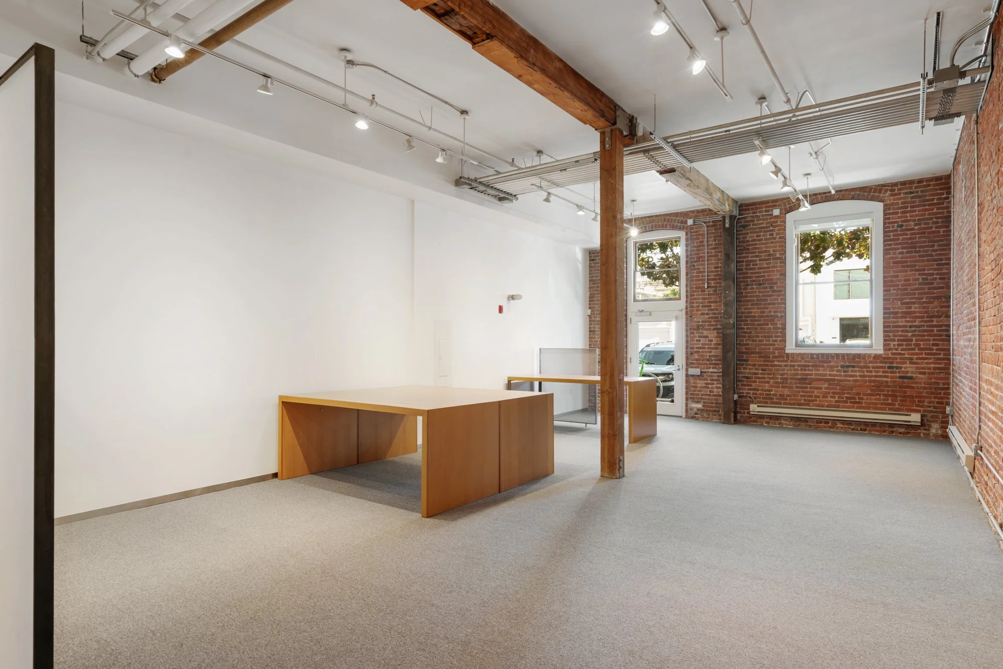 Empty commercial or office space with exposed brick walls, large windows, and a gray carpeted floor. There are wooden desks, an open ceiling with track lighting, exposed pipes, and a wooden support beam.