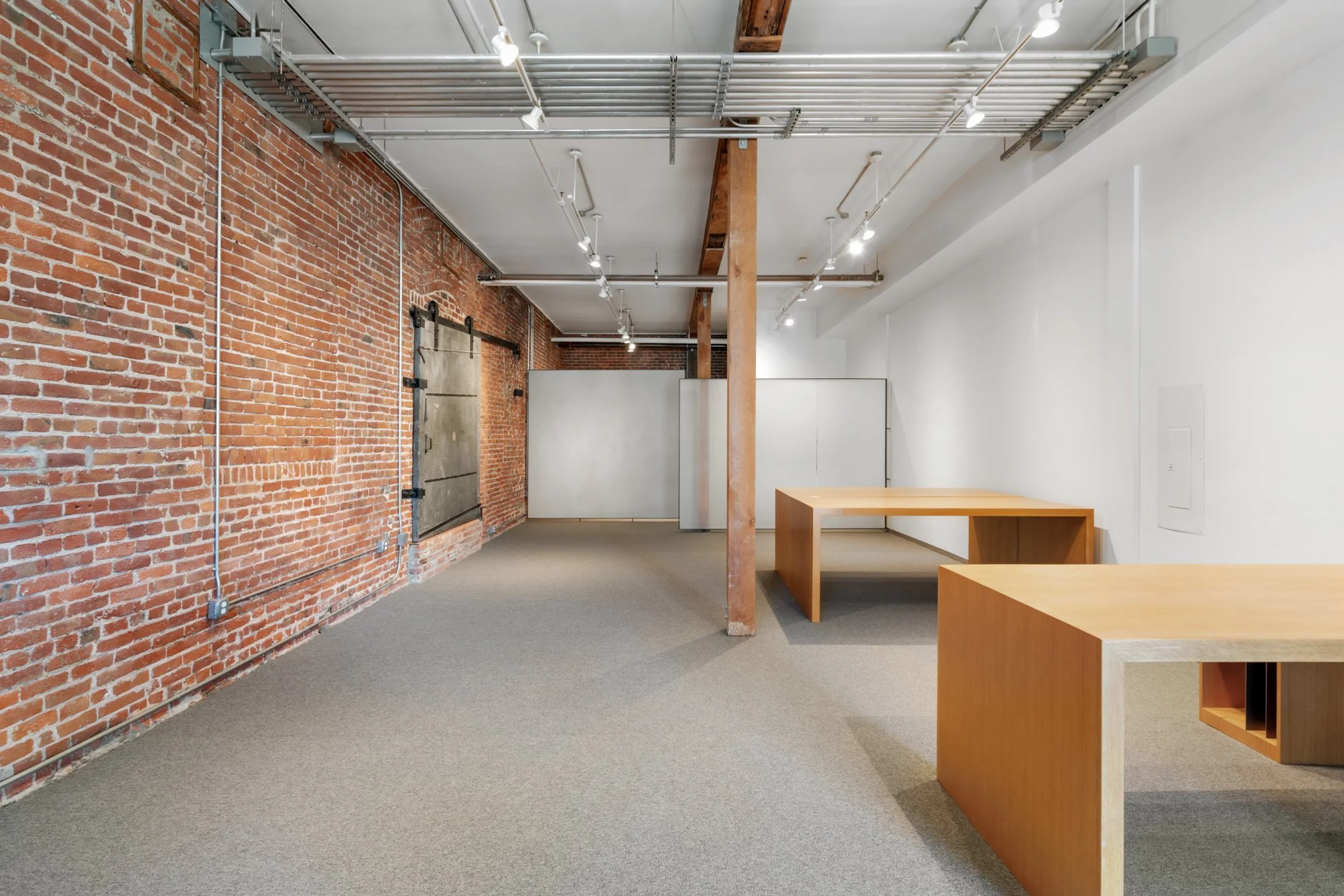 Empty office space with exposed brick walls, white painted walls, wooden desks, and track lighting fixtures.