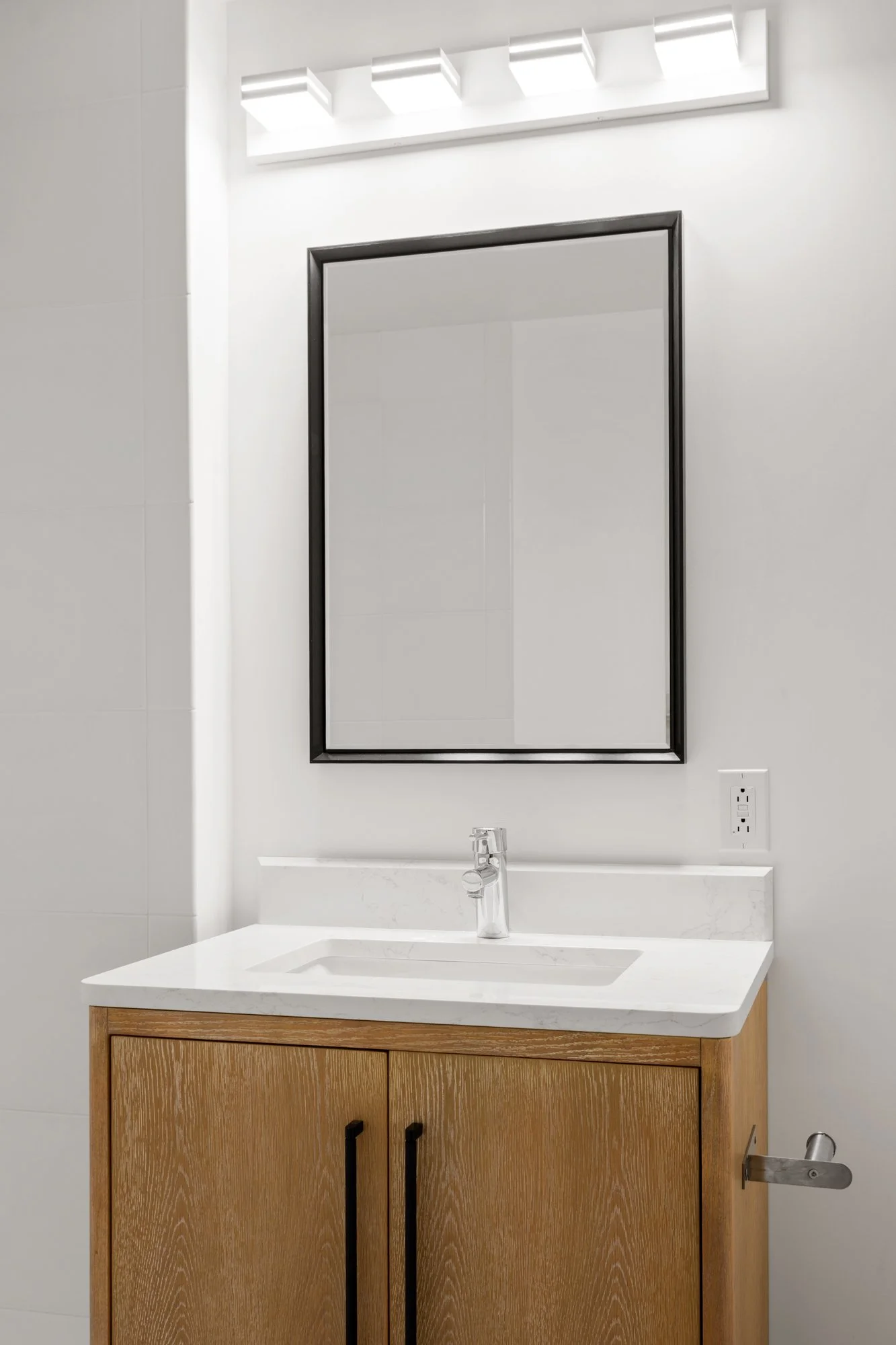 Modern bathroom vanity with a wooden cabinet, white countertop, and a large rectangular mirror with black frame, illuminated by overhead lighting