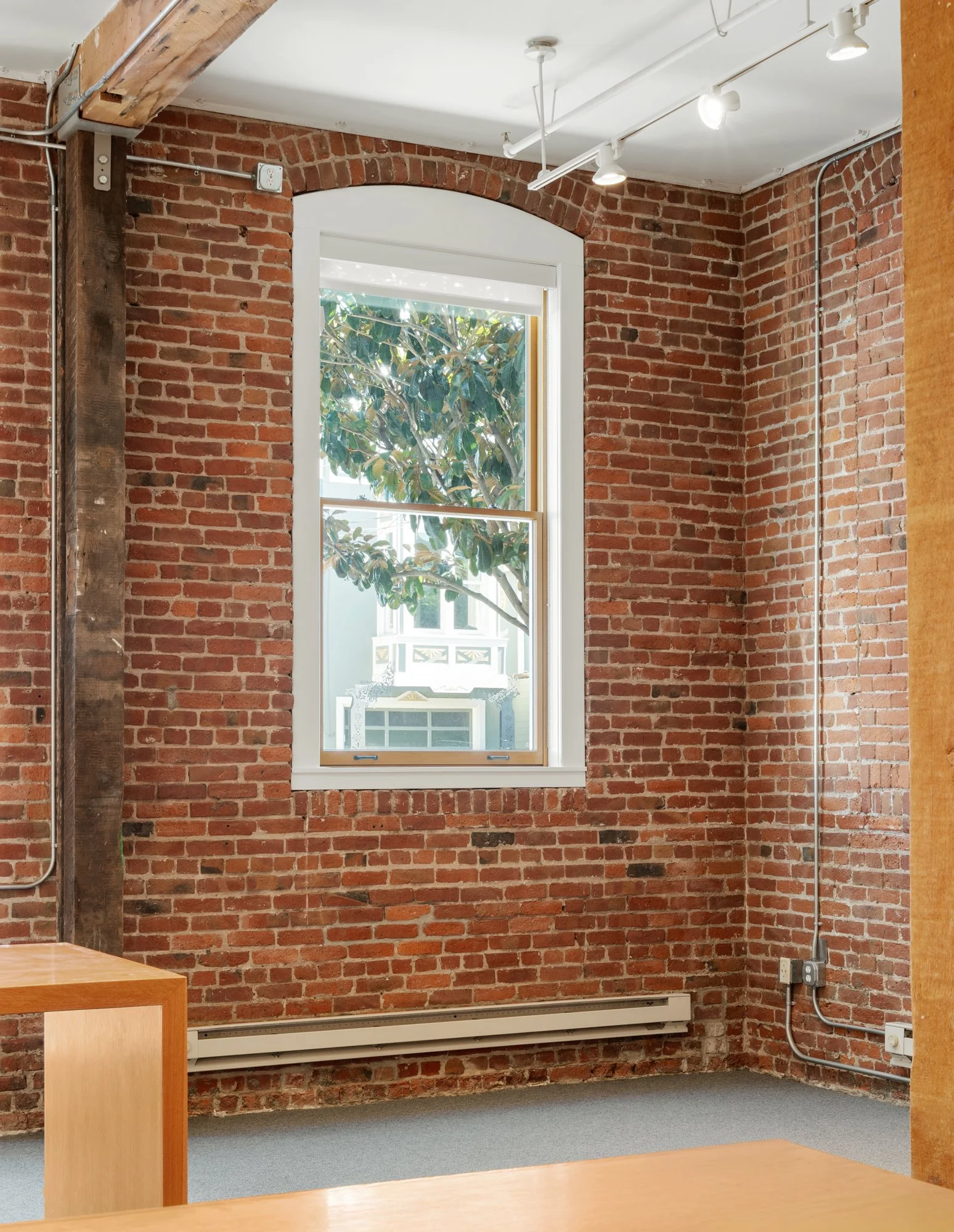 A brick-walled room with a window showing a view of a tree outside. The room has track lighting on the ceiling and exposed electrical conduit along the walls.