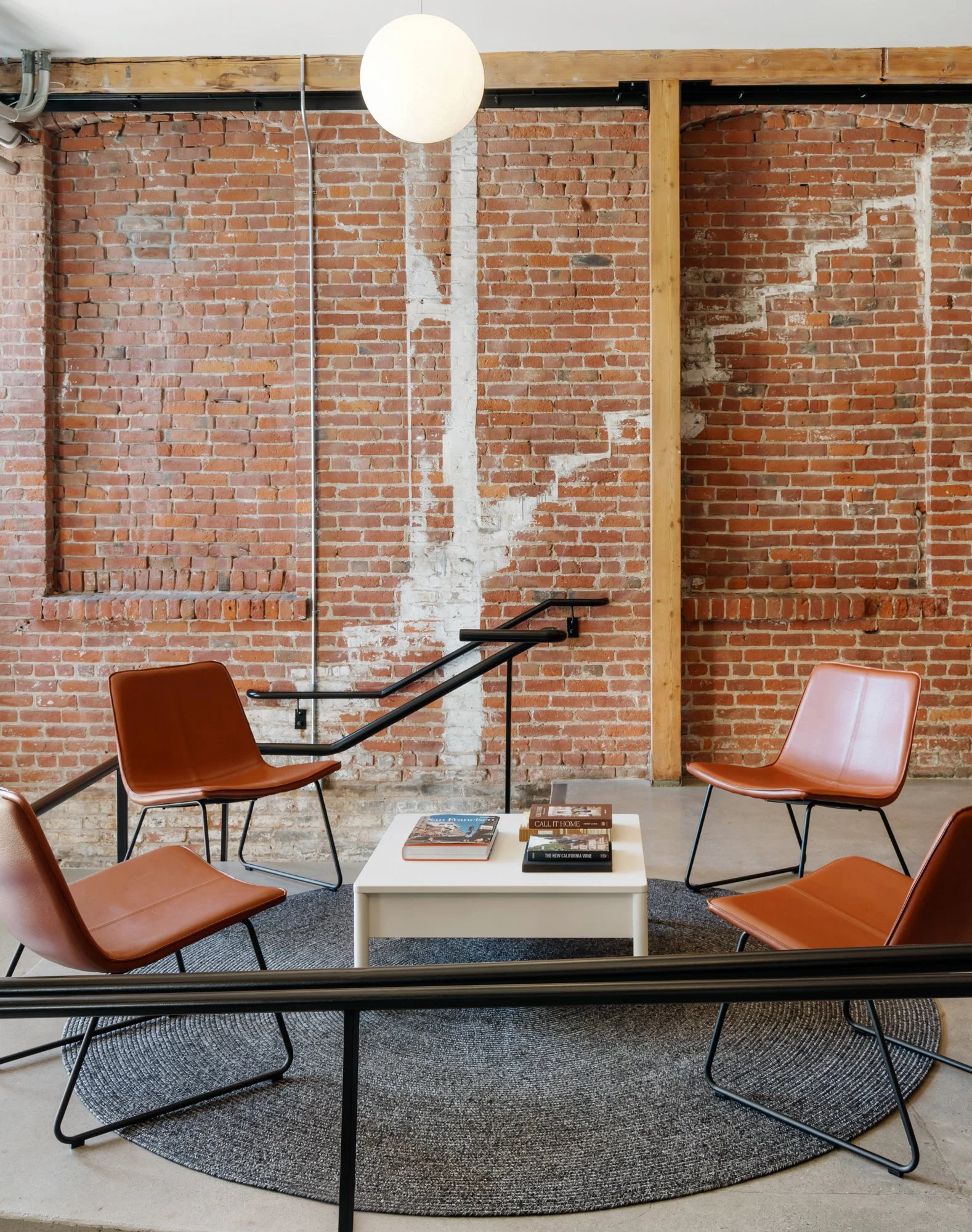 Modern lounge area with four brown leather chairs arranged in a circular pattern on a dark round rug, a white coffee table with magazines, exposed brick wall with a mix of repaired and unfinished bricks, and a hanging white round light fixture.
