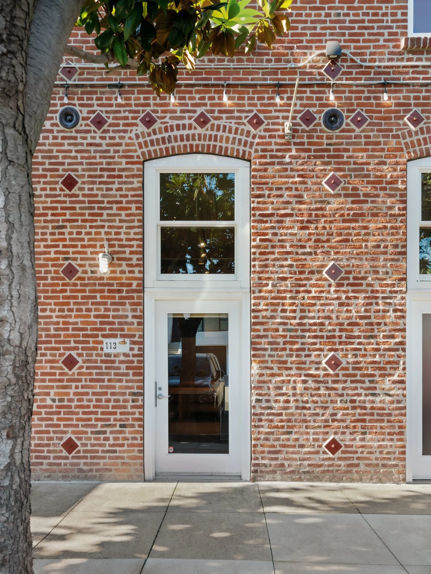 The image shows a brick building facade with a door and window, decorated with small brown tiles in diamond and square shapes, and string lights hanging across the top. There is an ID plaque with the number 113 next to the door.