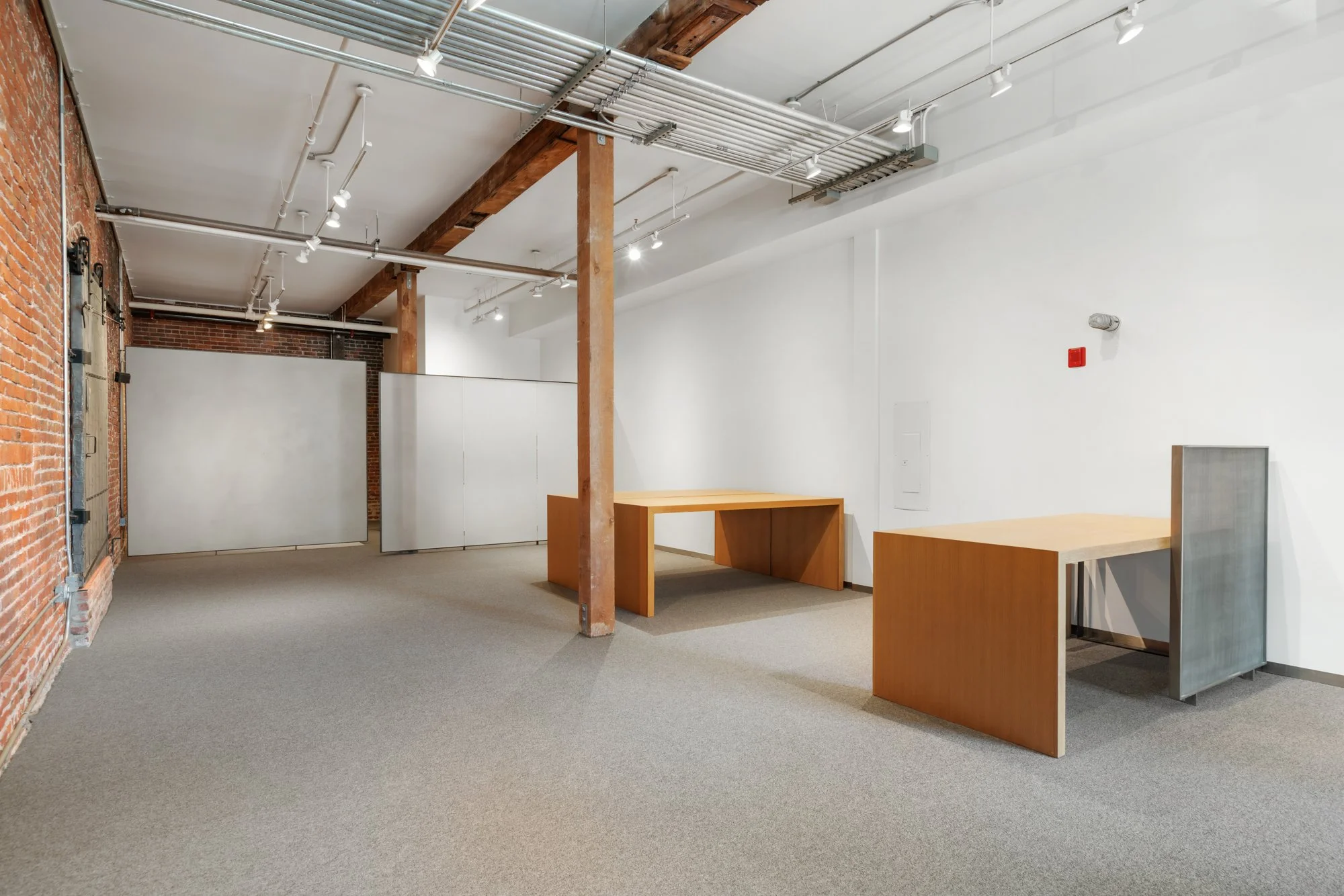 Empty art gallery interior with exposed brick wall, wooden beams, white walls, and display tables.