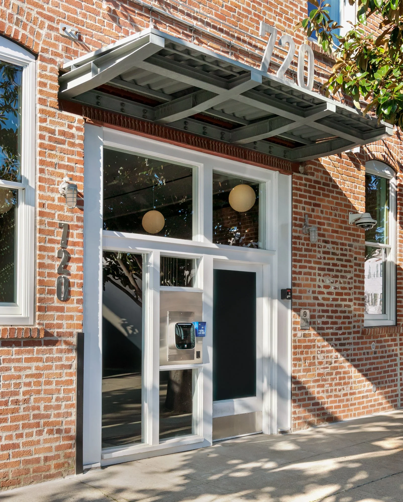 Brick building with modern entrance, including a door, large windows, an ATM, and the street number 720. A metal awning with the number 120 above the doorway, and two spherical lights inside visible through the windows. Sidewalk and shadows from near