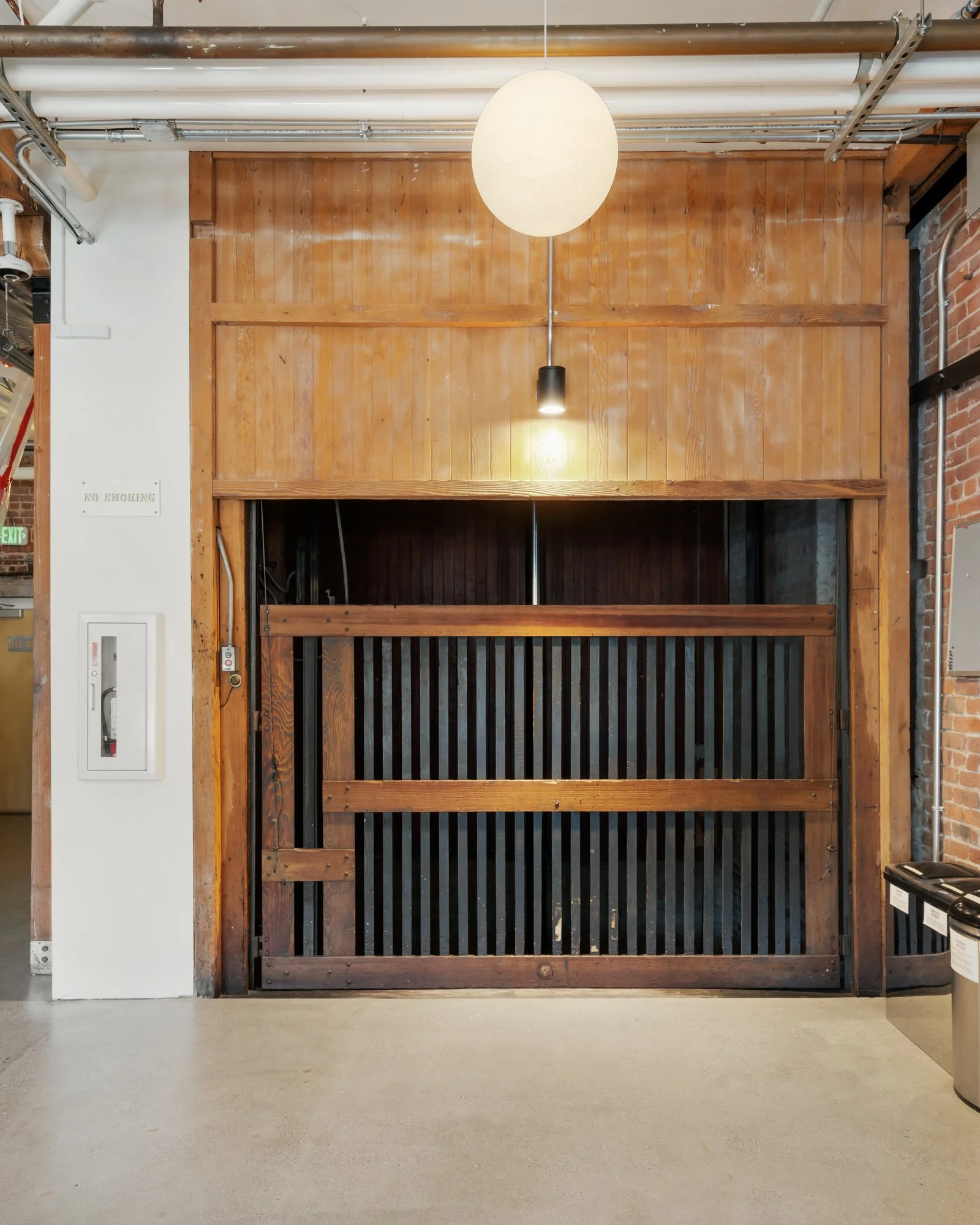 Warehouse or industrial-style wooden garage door with black vertical slats, surrounded by wood paneling and brick wall, lit by a hanging spherical light and a smaller spotlight.