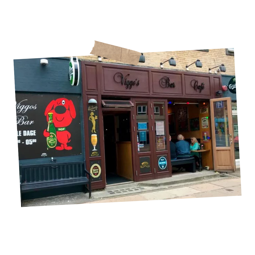Udefra viser et pub- eller barfacade med navnet 'Viggo's Bar & Café', der har en åben dør, hvor tre personer sidder og taler ved et bord. Der er plakater, skilte og enmalet logo ved indgangen.