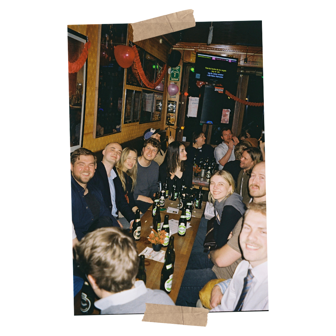 En gruppe unge mennesker sidder sammen ved et langbord i en bar med drikkevarer, latter og snak, festlige dekorationer som balloner og serpentiner synlige.