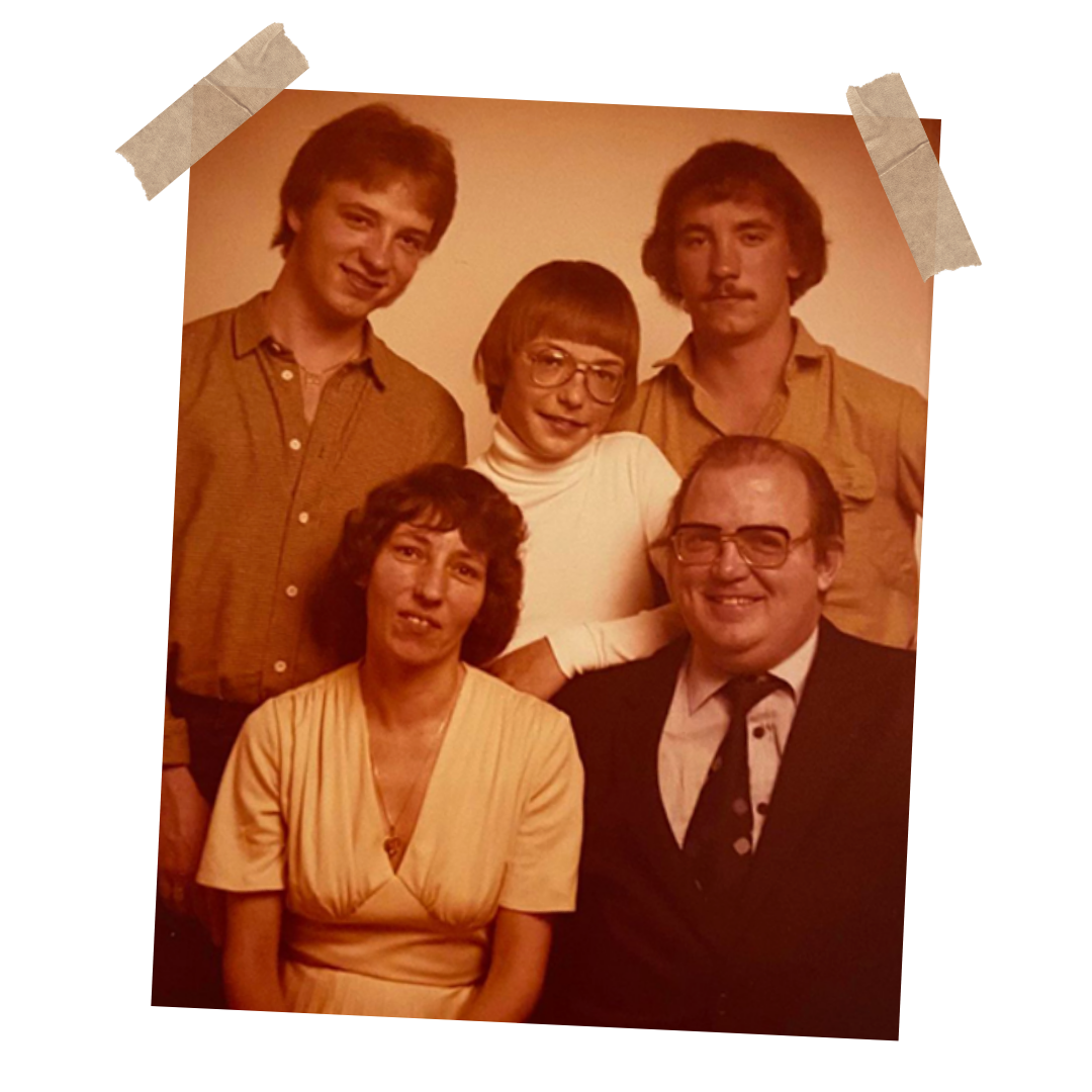 Familiebillede med seks personer, tre unge mænd, to kvinder og en teenage pige, alle iført 1970'er tøj, samlet i en gruppe for et portræt.