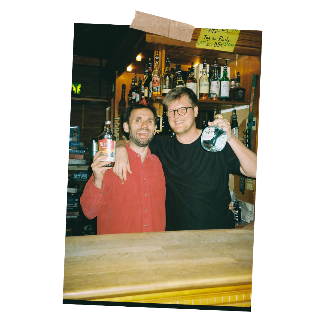To mænd, der holder alkoholholdige drikkevarer, smiler foran en bar med forskellige flasker alkohol i baggrunden.