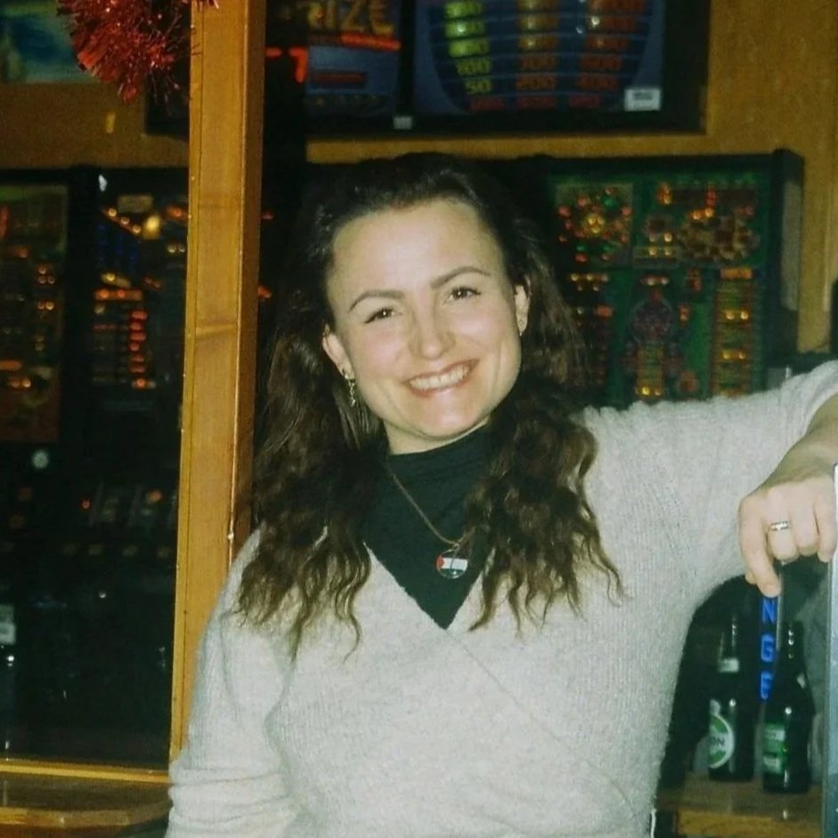 Et smilende kvindemenneske med langt, krøllet brunt hår, iført en sort top under en lysegrå sweater, står i en bar eller casino med spilleautomater i baggrunden