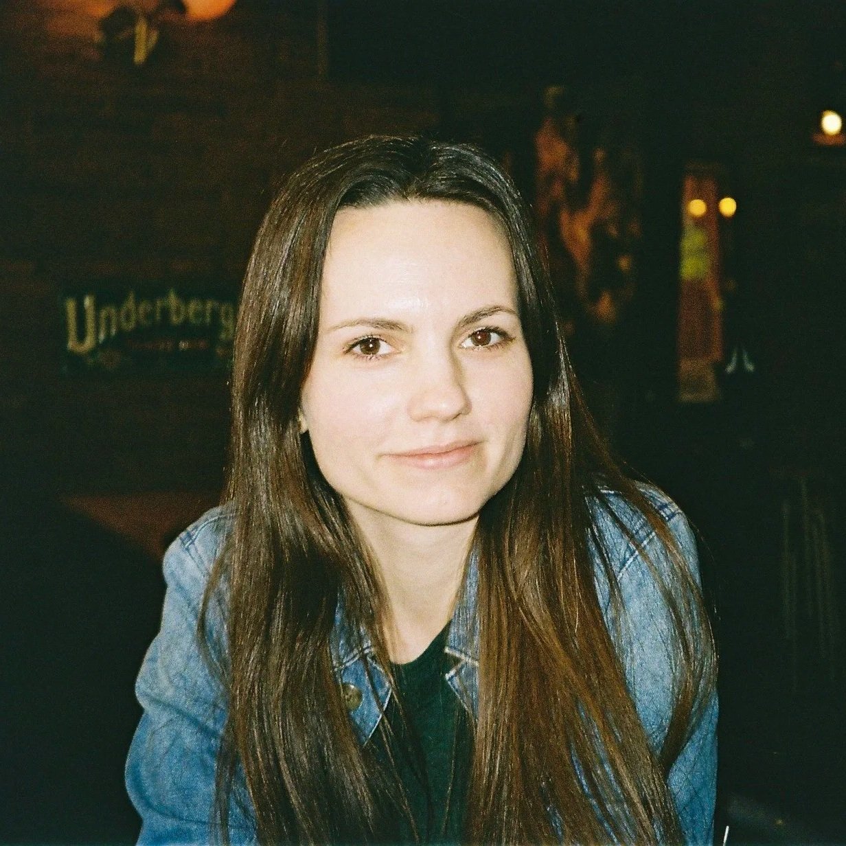 Et portræt af en ung kvinde med langt brunt hår, der sidder i en restaurant eller bar med svagt belysning.