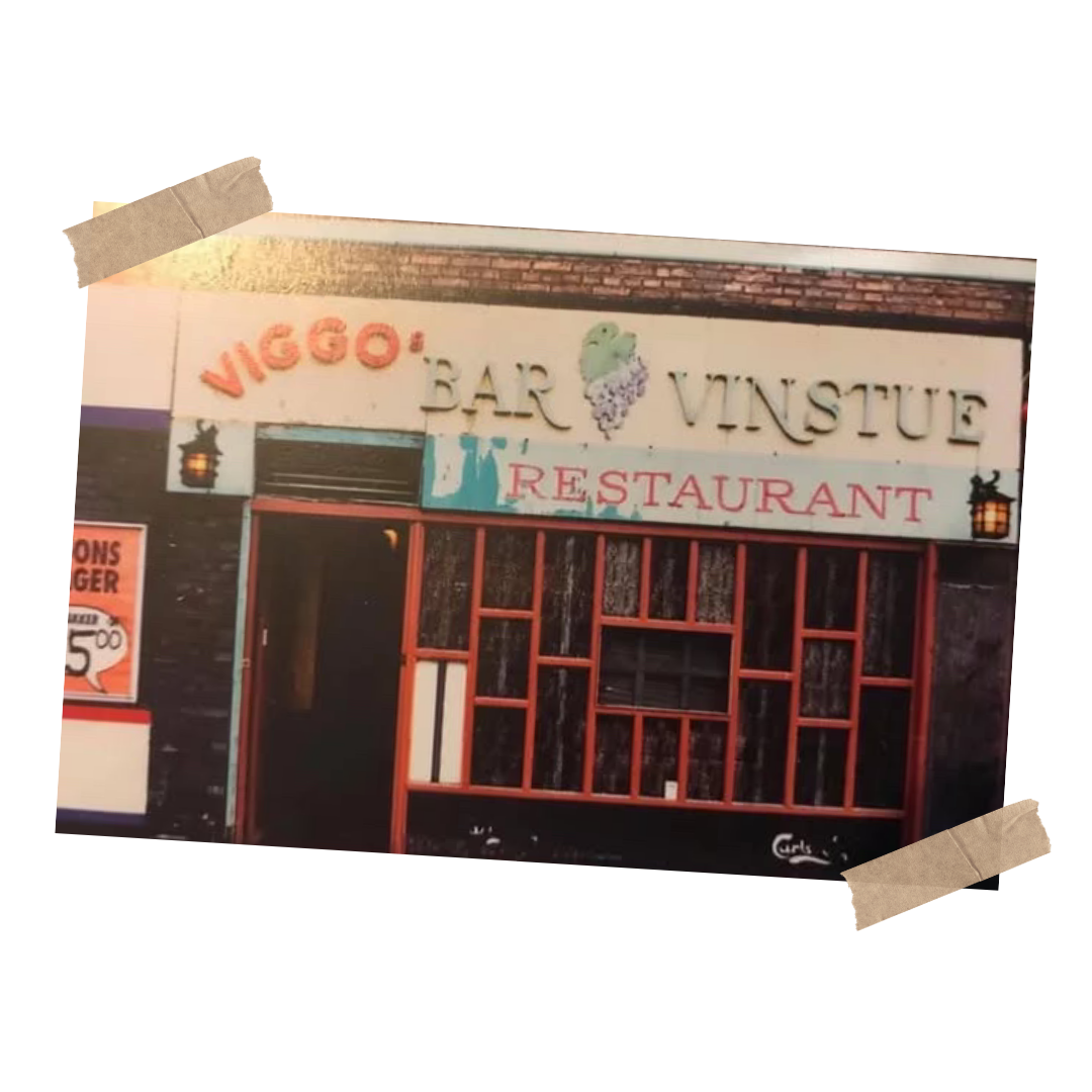 En restaurantfacade med et skilt, der siger 'VIGO: BAR VINSTUE RESTAURANT' og et billede af en vindrueklase. Der er også tilhørende lamper og nye tømmerlister.