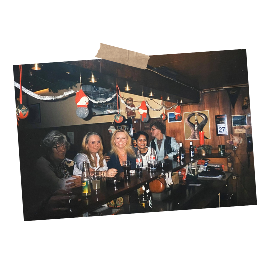 Fem personer sidder ved en bar med juledekorationer, herunder nissehuer og julepynt, og hænger ud i en hyggelig indretning.