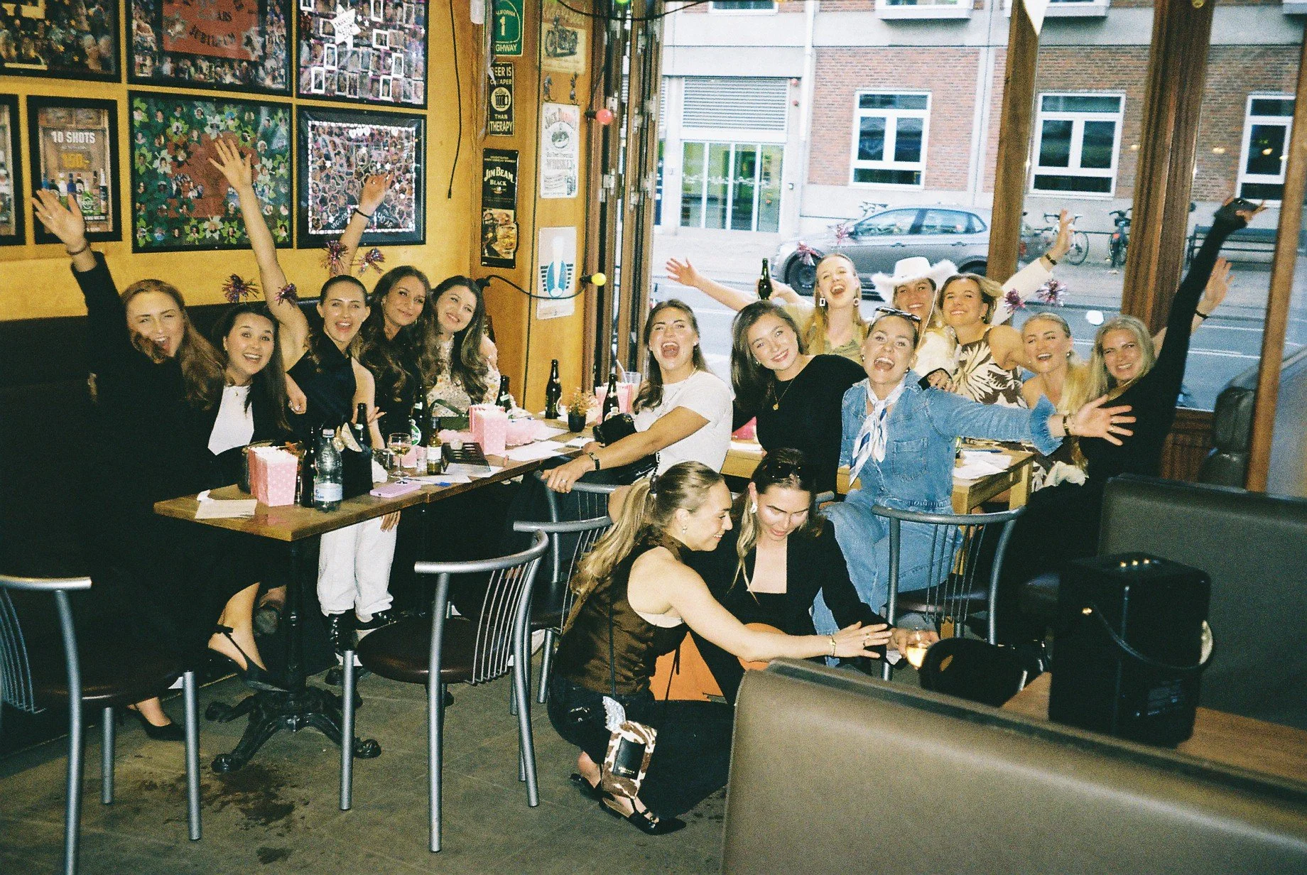 En gruppe kvinder, der sidder og står rundt om et bord i en restaurant, smiler og fejrer, med nogle af dem løftet i armene og nogle med festlige accessories. Der er drikkevarer og små gaver på bordet, og et vindue med udgang til en gade udenfor.